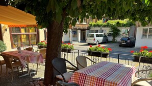 An outdoor seating area with wicker chairs and tables covered with checkered tablecloths, surrounded by potted plants and a tree. - Hotel Bären - The Bear Inn (Wilderswil)