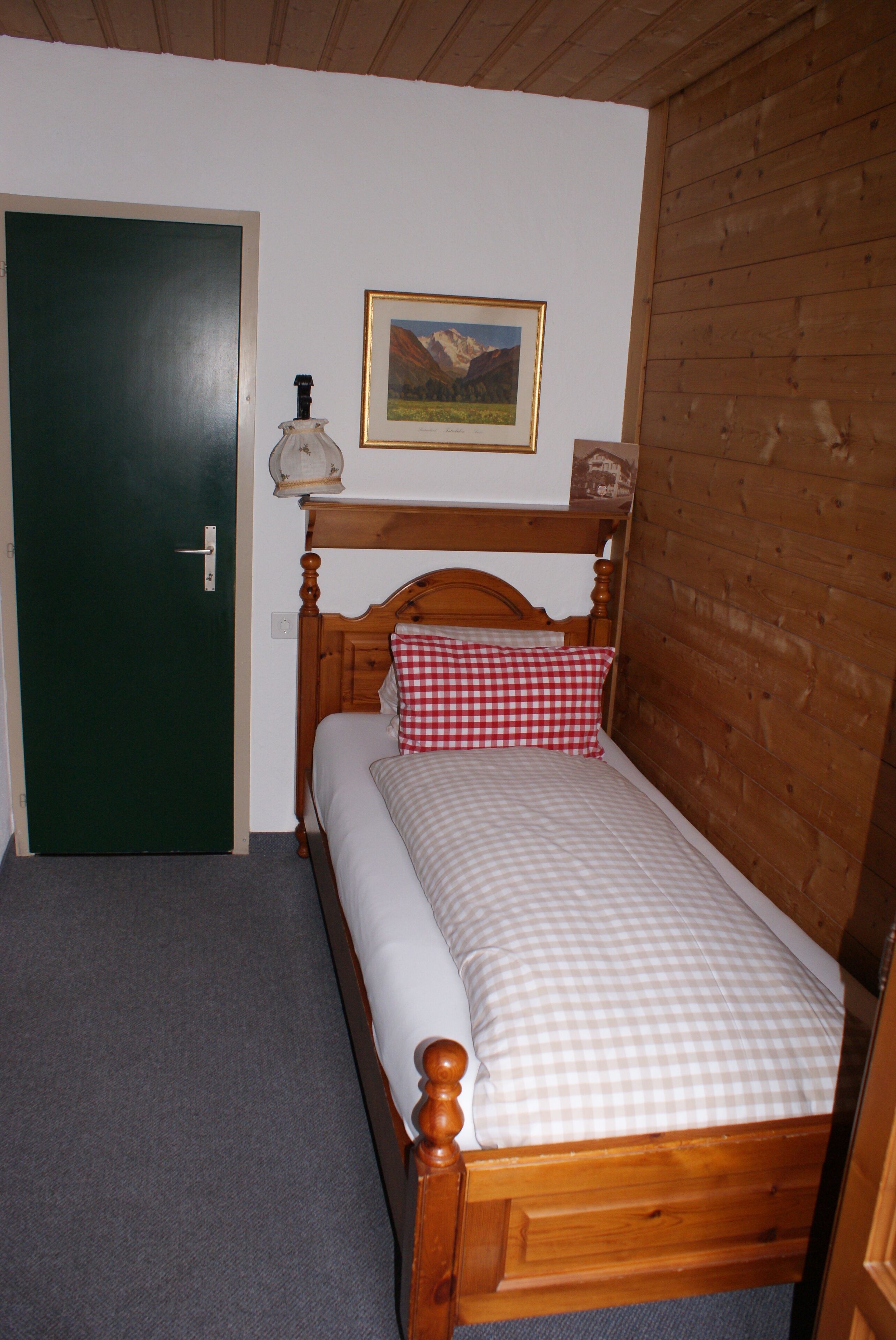 Ein Zimmer mit einem Einzelbett, einem Holzkopfende, einer grünen Tür, einem gerahmten Bild an der Wand und einem kleinen Tisch mit einer Lampe.