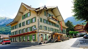 Un hotel de varios pisos con contraventanas verdes, un letrero que dice 'HOTEL BAREN' y la silueta de una montaña al fondo.