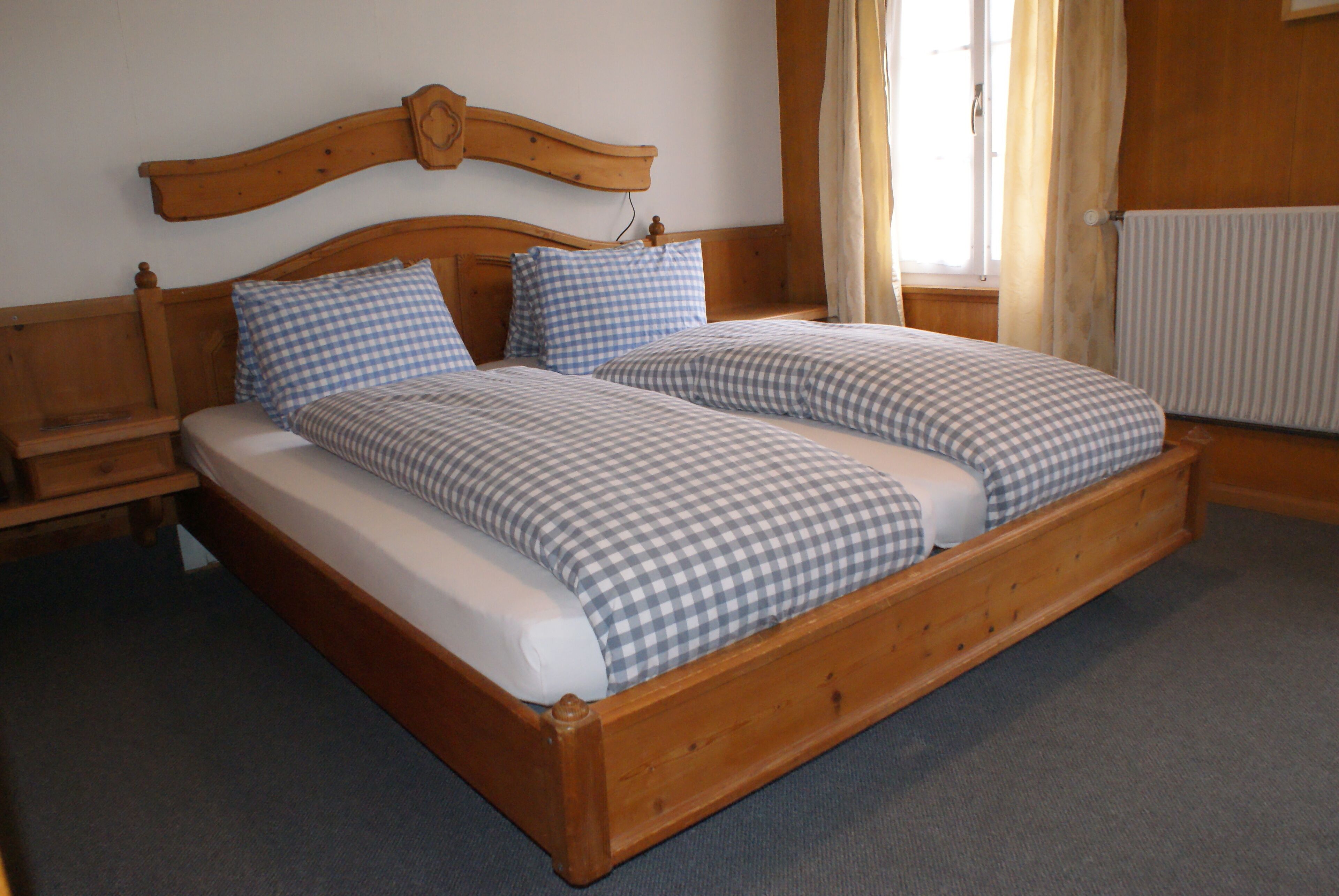 A double bed with a wooden frame and checkered bedding. There is a nightstand on the left, a window with curtains, and a radiator on the right.