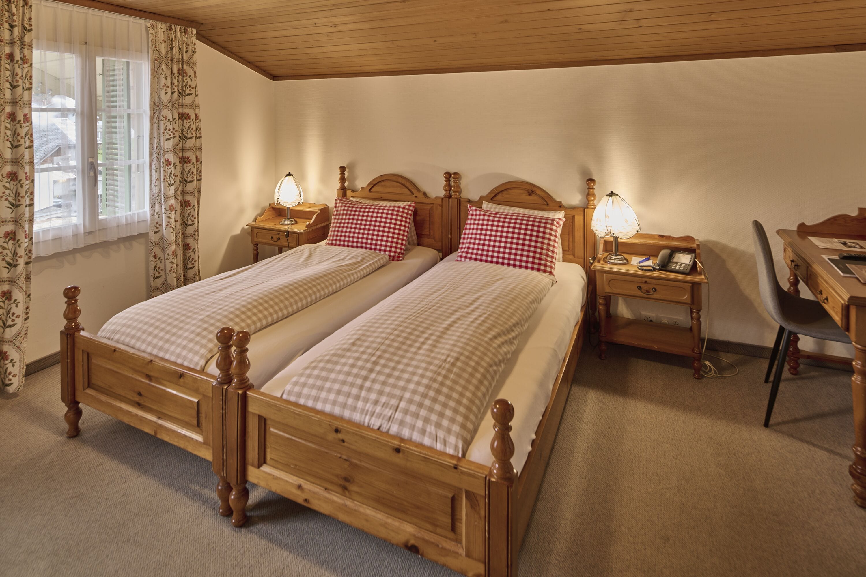 Ein Doppelbett aus Holz mit karierter Bettwäsche, zwei Nachttische mit Lampen, ein Schreibtisch mit Stuhl und ein Fenster mit Vorhängen.