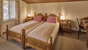 A double wooden bed with checkered bedding, two bedside tables with lamps, a desk with a chair, and a window with curtains.