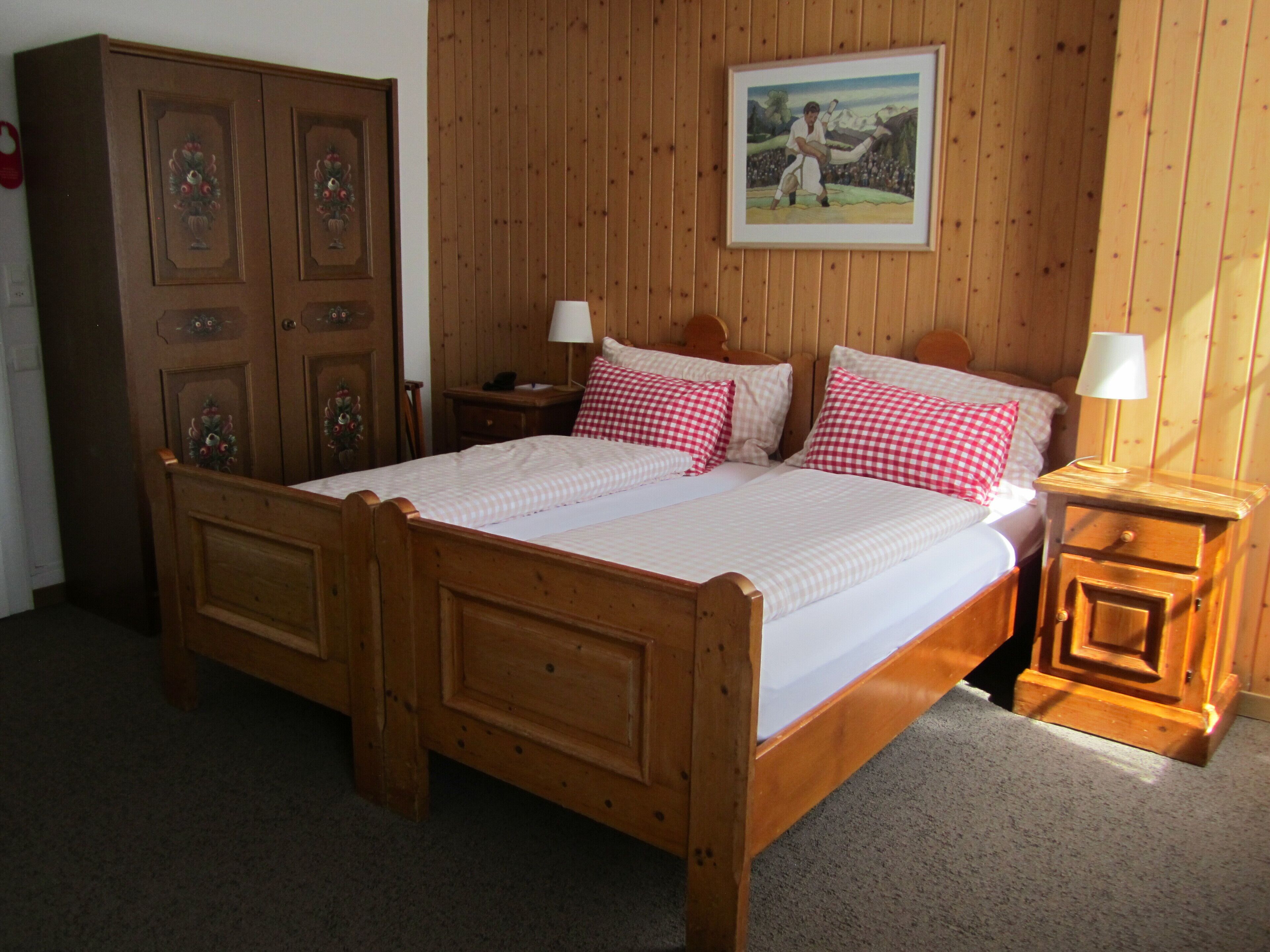 Un lit à baldaquin en bois, recouvert d’une courtepointe blanche et agrémenté d’oreillers à carreaux rouges et blancs. De chaque côté du lit, on trouve une table de chevet en bois sur laquelle est posée une lampe. Une armoire en bois, ornée de panneaux décoratifs, se trouve à gauche du lit.