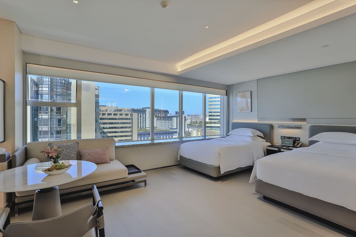 A modern hotel room with a large window, two beds, a sofa, a dining table, and a small kitchen area.
