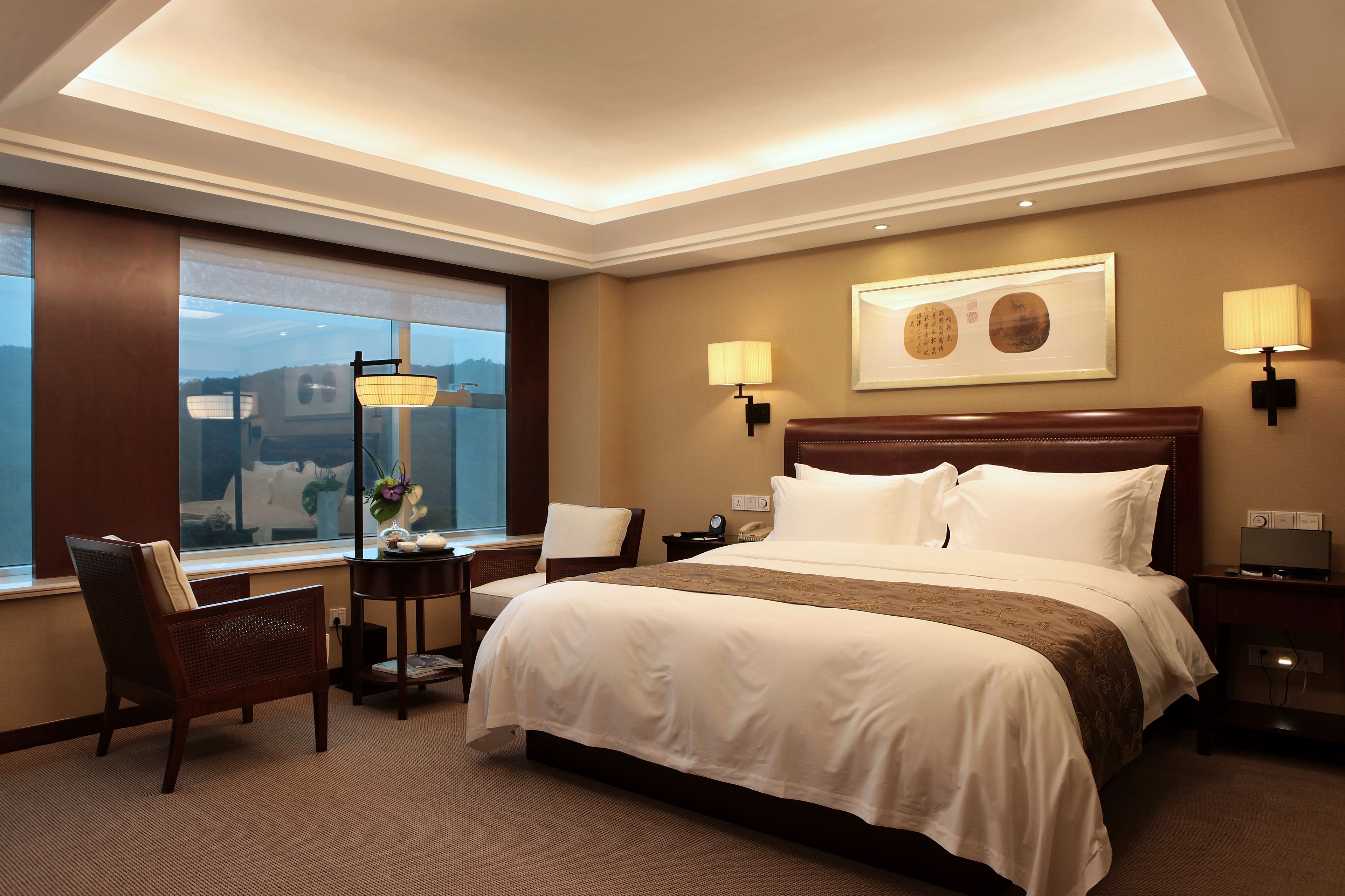 Une chambre d’hôtel avec un grand lit, deux tables de chevet, une chaise, un bureau avec une lampe et une fenêtre donnant sur un beau paysage.