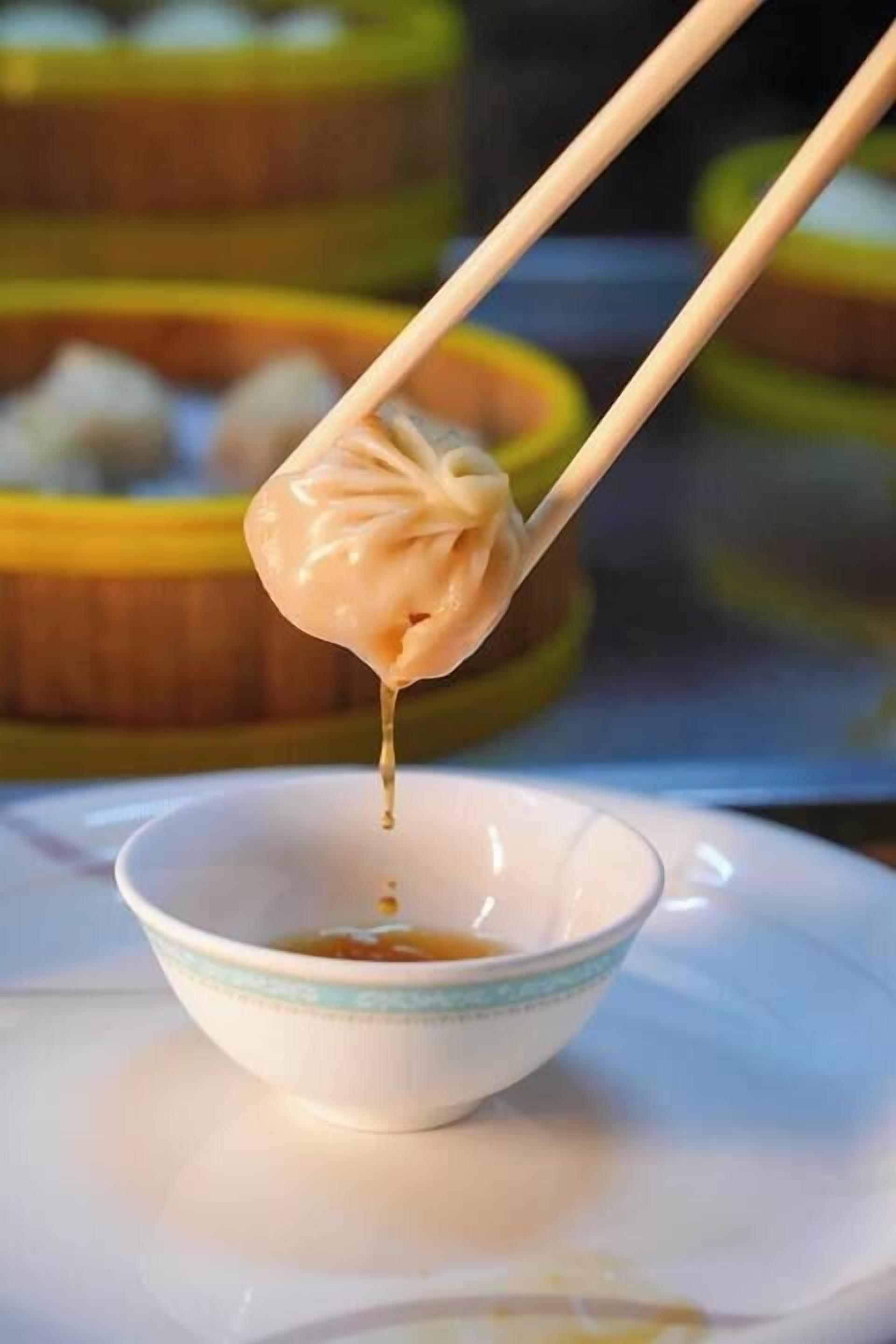A bowl of soup with chopsticks lifting a dumpling, with a blurred background of similar bowls.