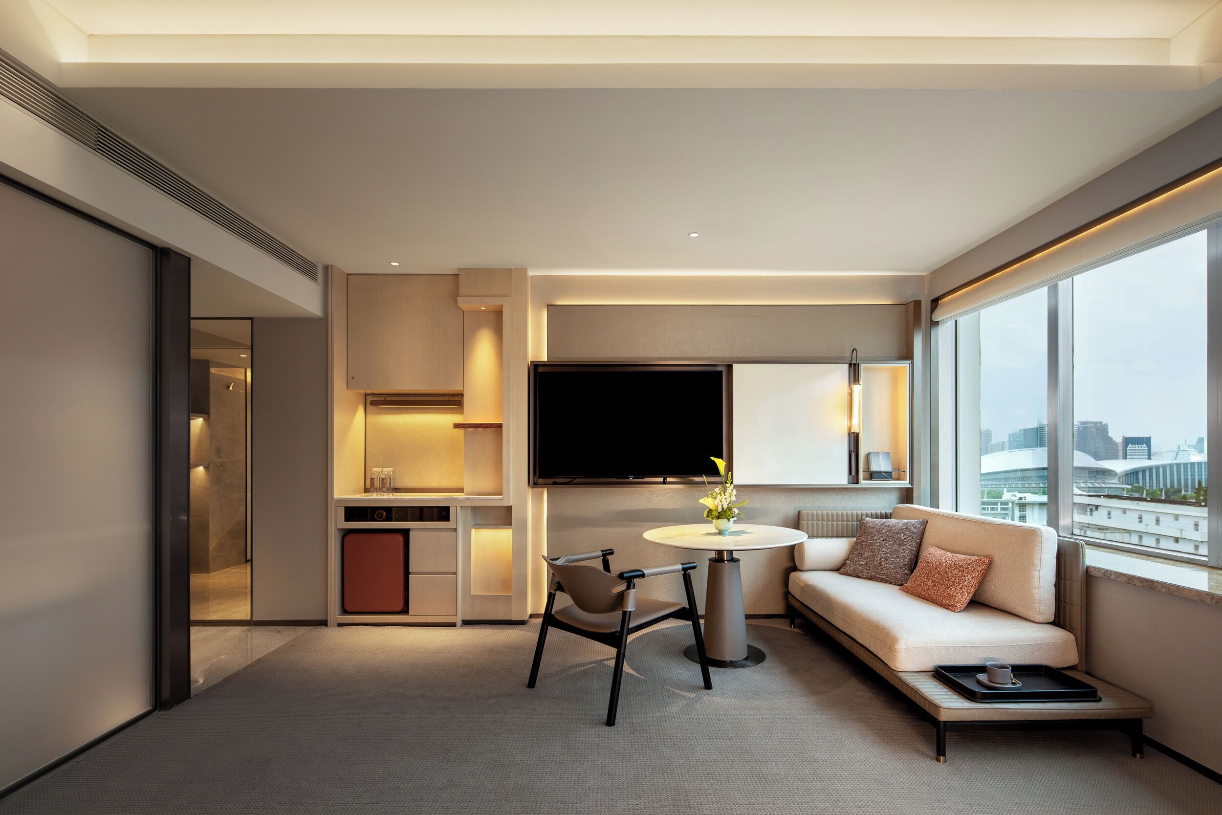 A modern hotel room with a sofa, a dining table, a TV, and a city view.