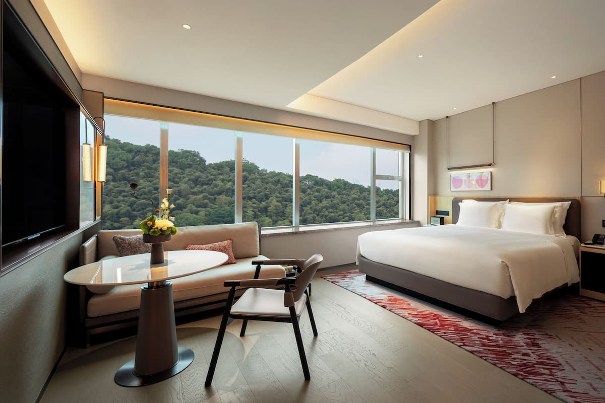 A modern hotel room with a large bed, a sofa, a dining table, and a view of a forested hill.