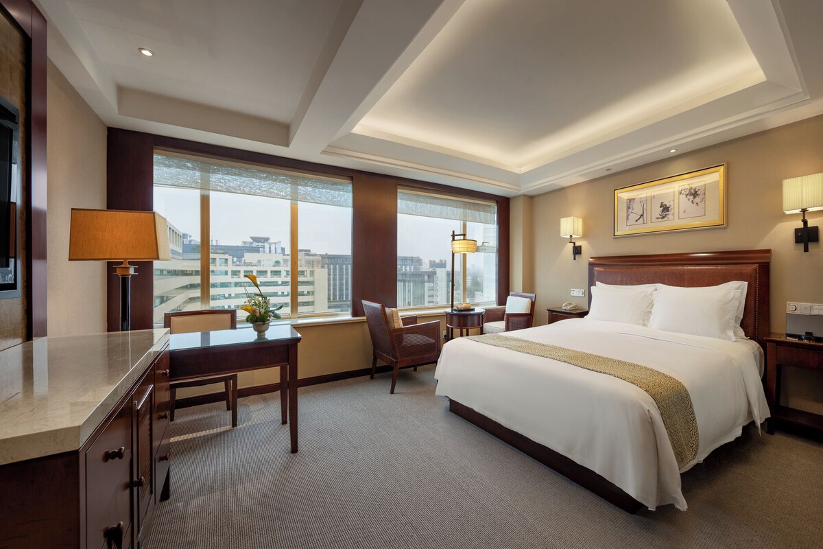 A hotel room with a large bed, a desk, a chair, and a TV. There are windows with a view of buildings outside.