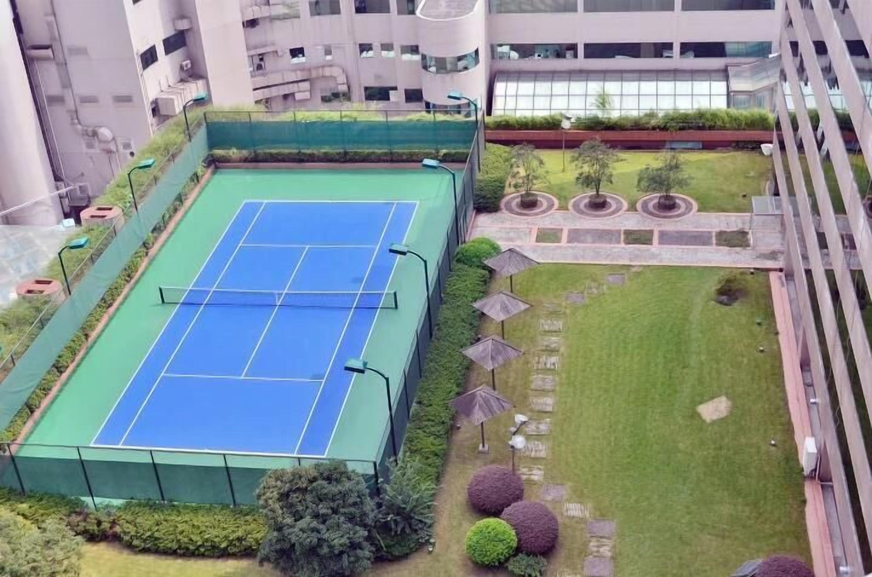 An outdoor tennis court surrounded by greenery and a building complex.