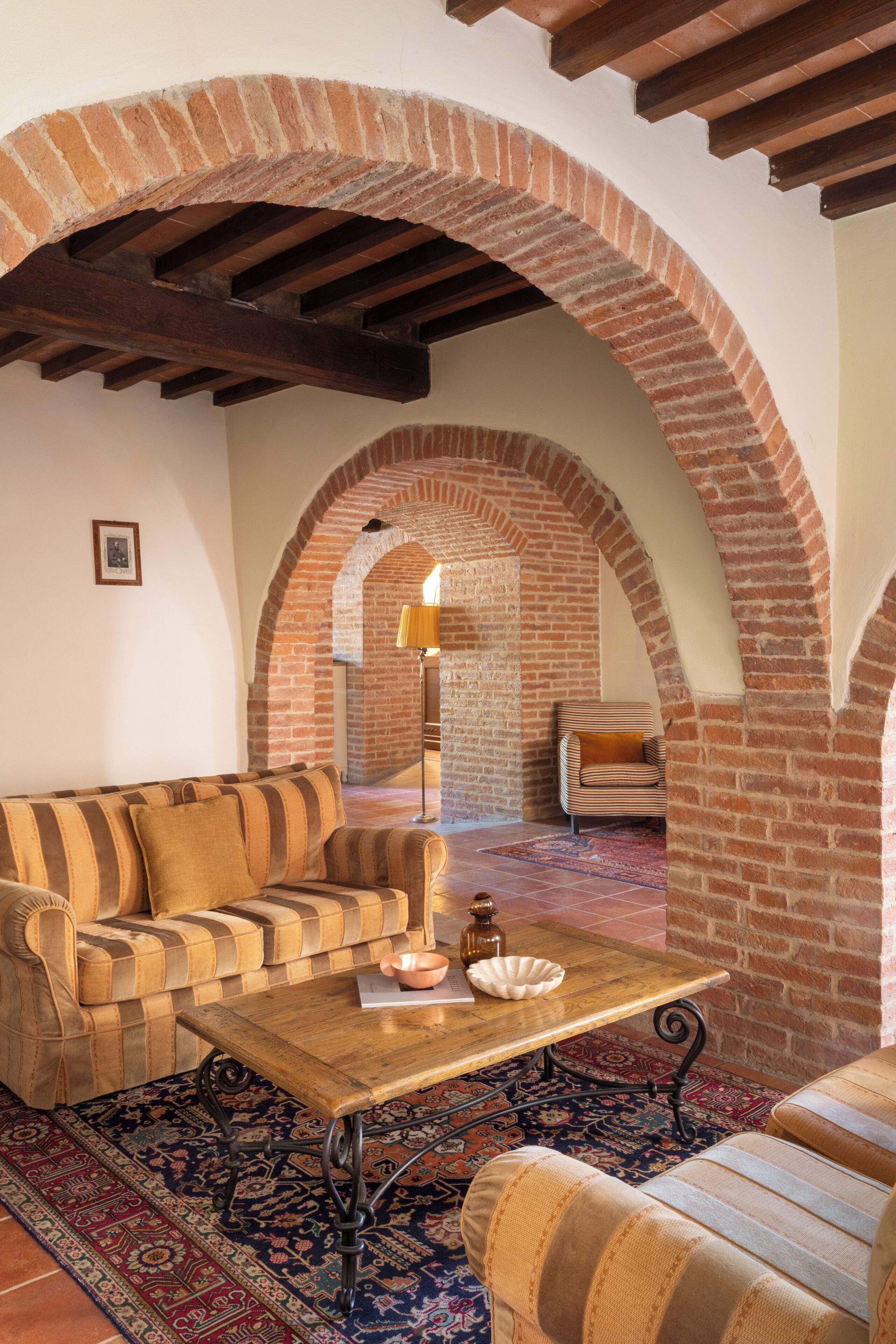 A cozy living room with a sofa, armchair, and coffee table, under a wooden ceiling and brick arches.
