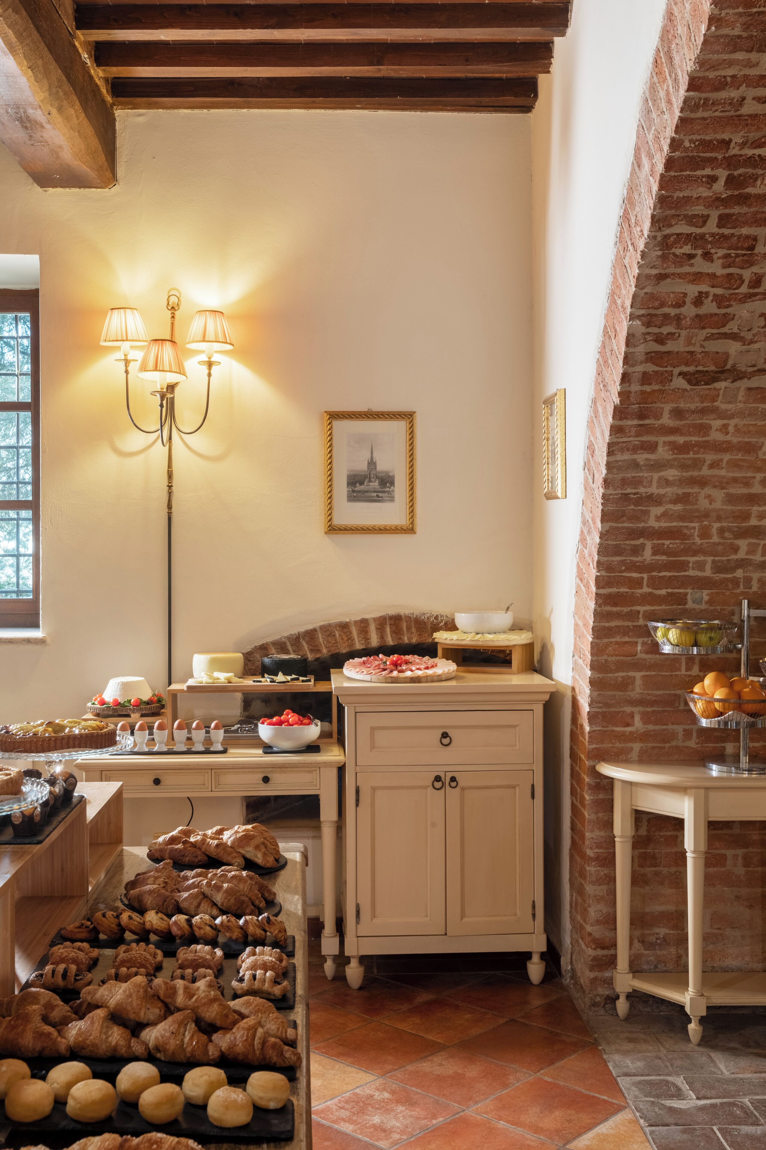 A kitchen with a brick wall, wooden beams, and a buffet table with pastries and a cake.