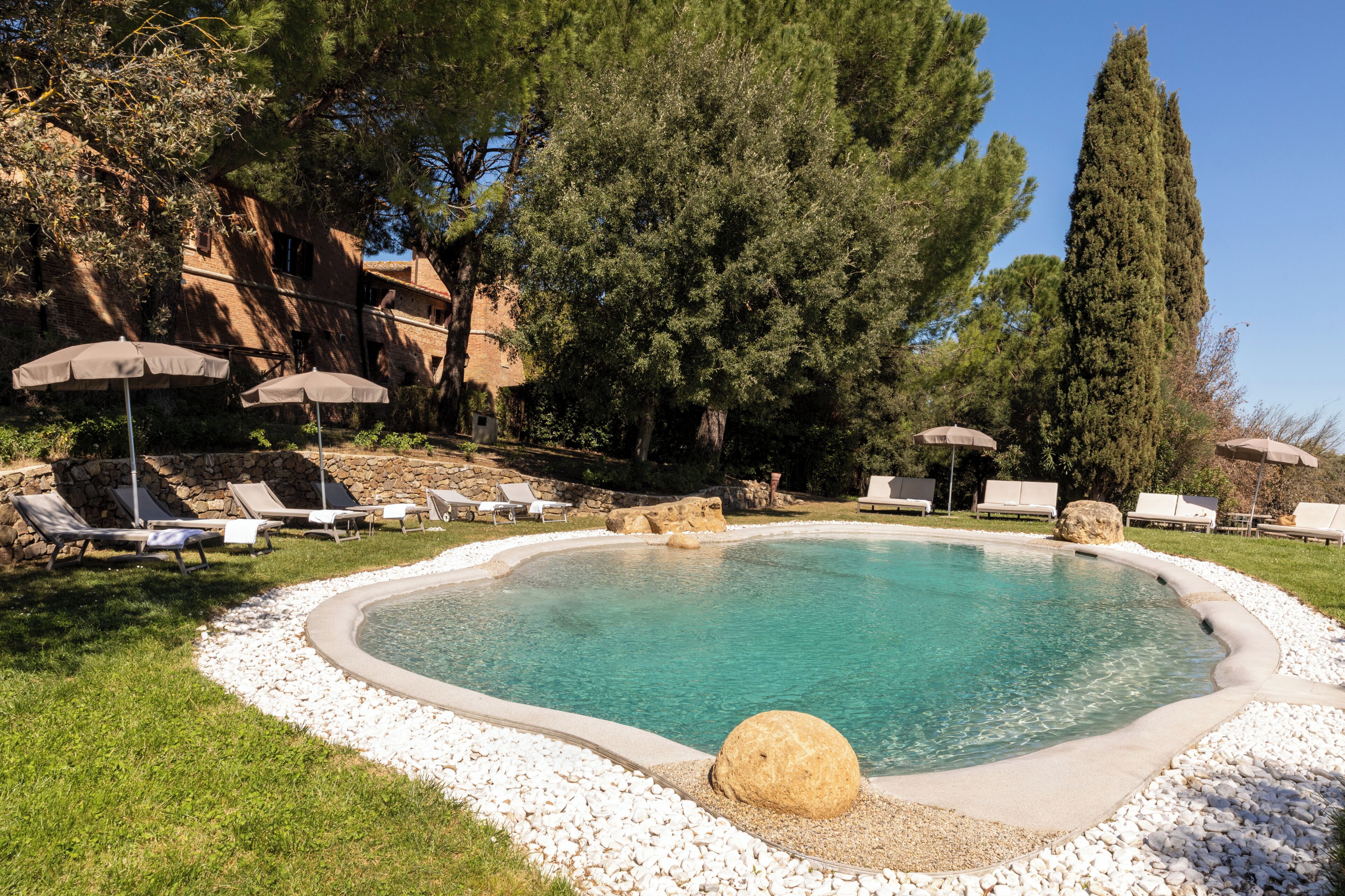 Une piscine entourée d’arbres et d’un mur de pierre, avec des chaises longues et des parasols à proximité.