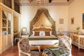 A bedroom with a four-poster bed, a chandelier, a desk with a chair, and a console table with a vase of flowers. - Castello di Leonina Relais (Asciano)