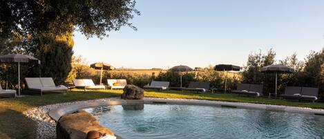 A pool area with lounge chairs and umbrellas, surrounded by rocks and a grassy area.