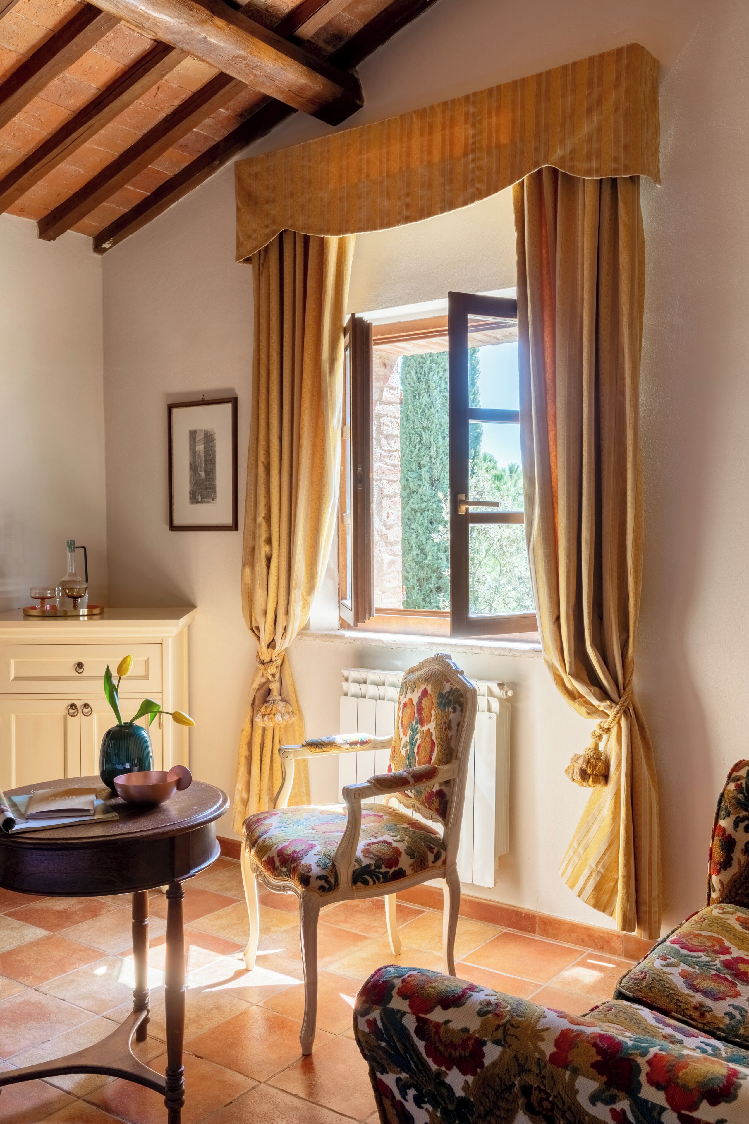 A room with a wooden ceiling, a window with curtains, a chair, a table, and a radiator.