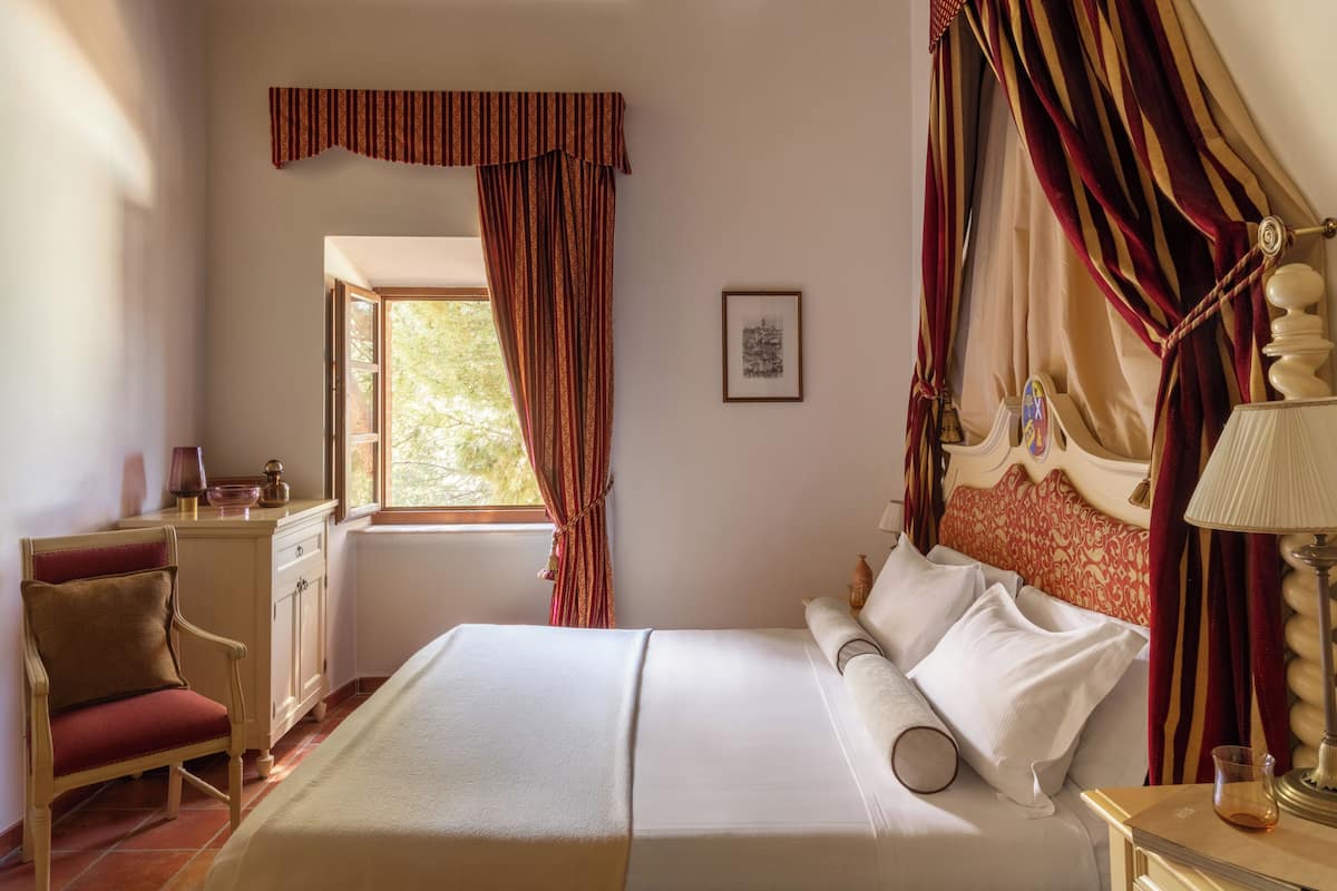 Un dormitorio con cama, una silla, mesita de noche, lámpara y ventana con cortinas.