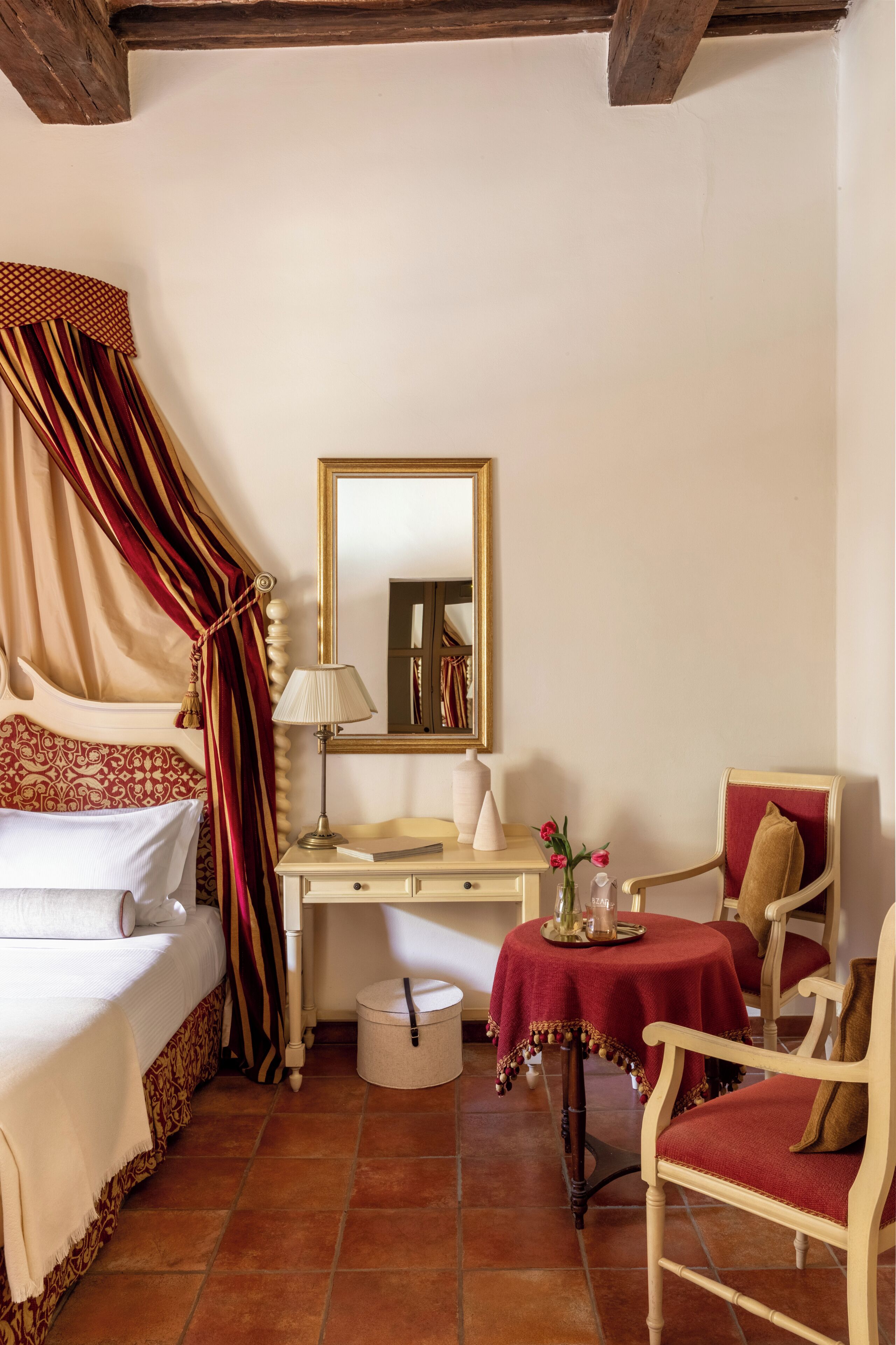 A bedroom with a bed, bedside table, lamp, mirror, chair, and a small round table with a red tablecloth.