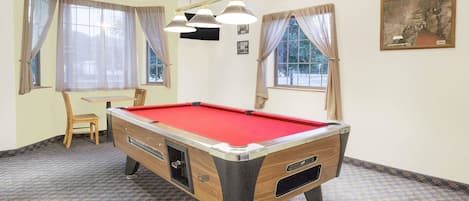 Een pooltafel in een kamer met een kamerbreed tapijt, een stoel en gordijnen voor de ramen.