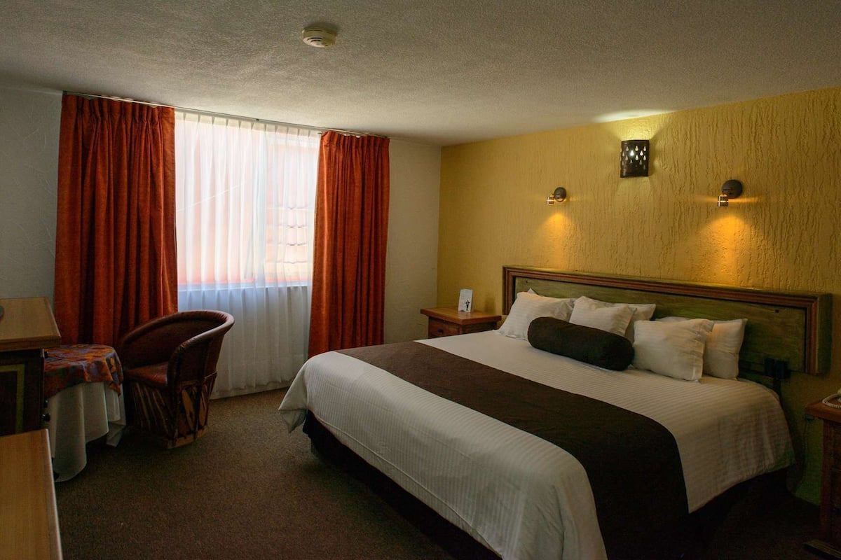 A hotel room with a large bed, a desk with a chair, a small table, and a window with curtains.