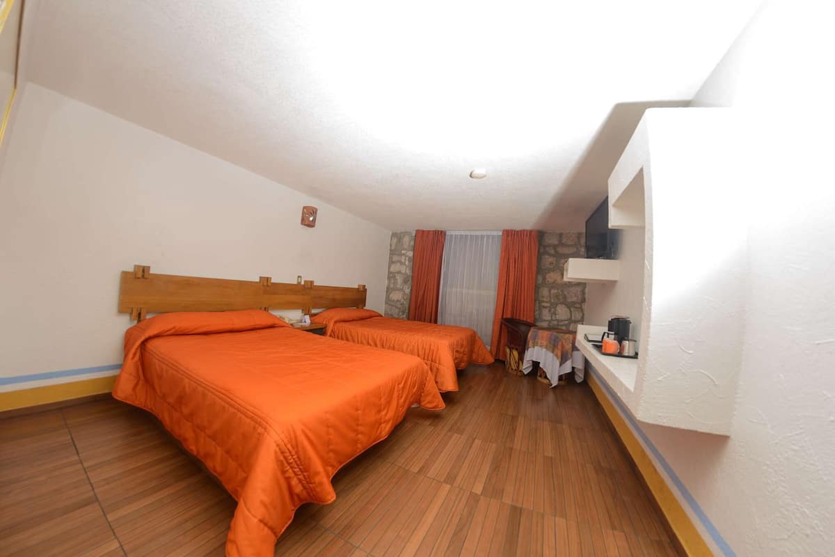 A hotel room with two beds, wooden headboards, a stone wall, a window with curtains, and a small table with a kettle.
