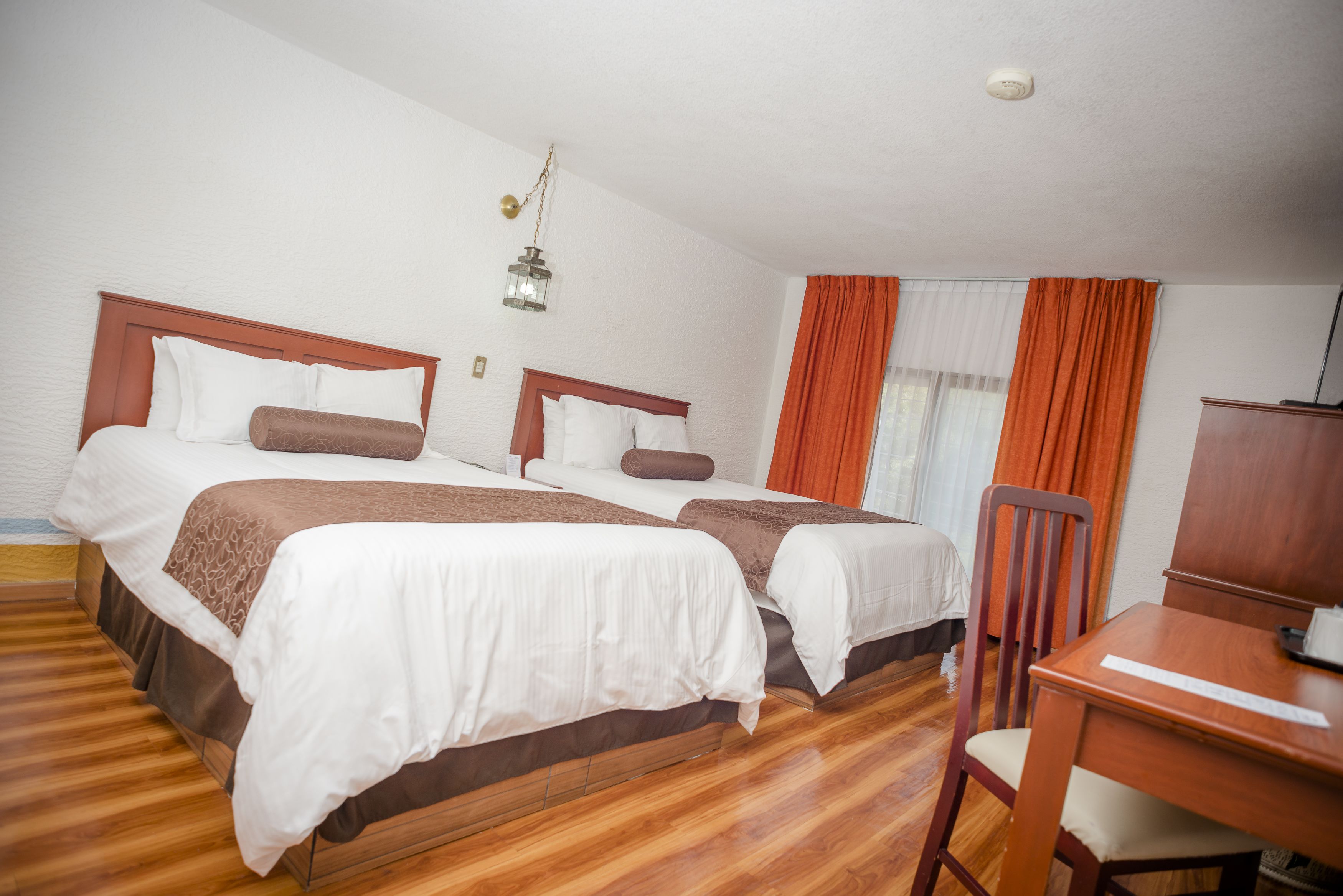 A hotel room with two beds, wooden flooring, a desk, and a chair.