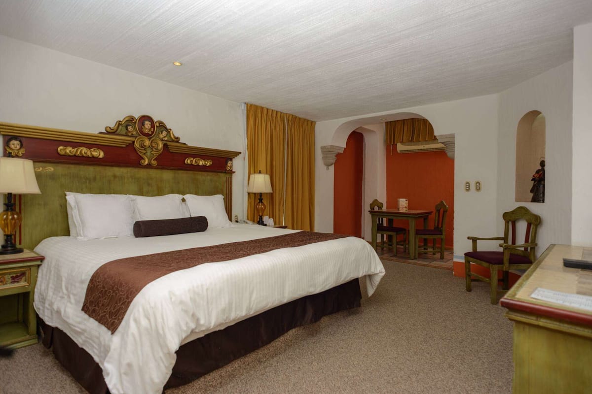 A hotel room with a large bed, a desk, and a chair. There is a small table with a lamp and a statue.
