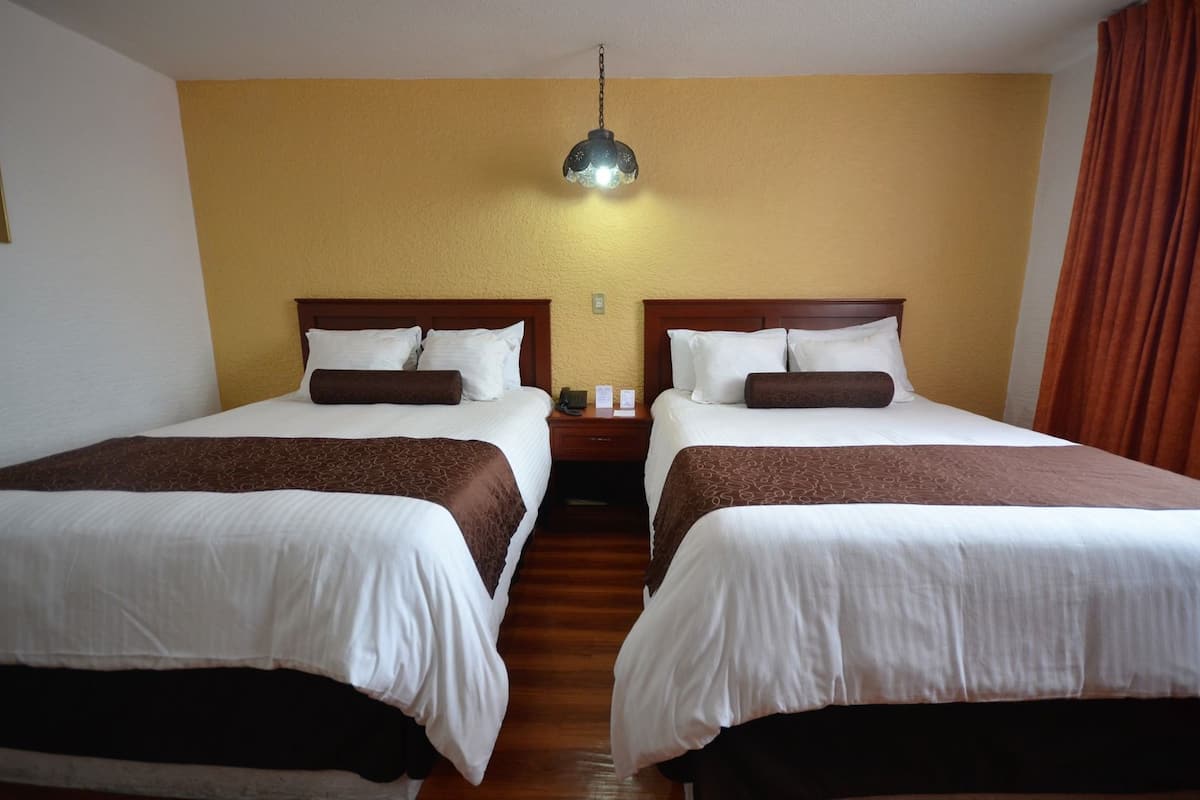 A hotel room with two beds, wooden headboards, a nightstand with a phone, and a hanging lamp.
