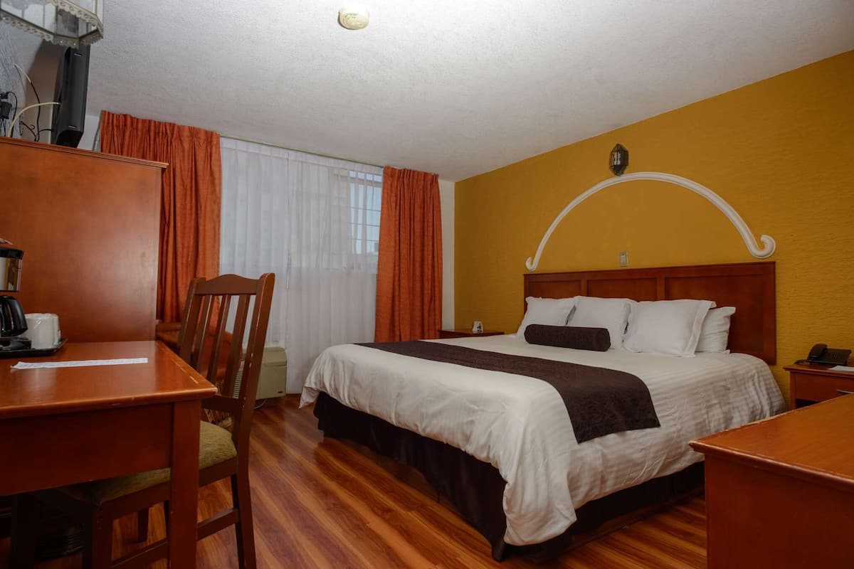 A hotel room with a wooden bed, a desk with a chair, a small table, and a window with curtains.