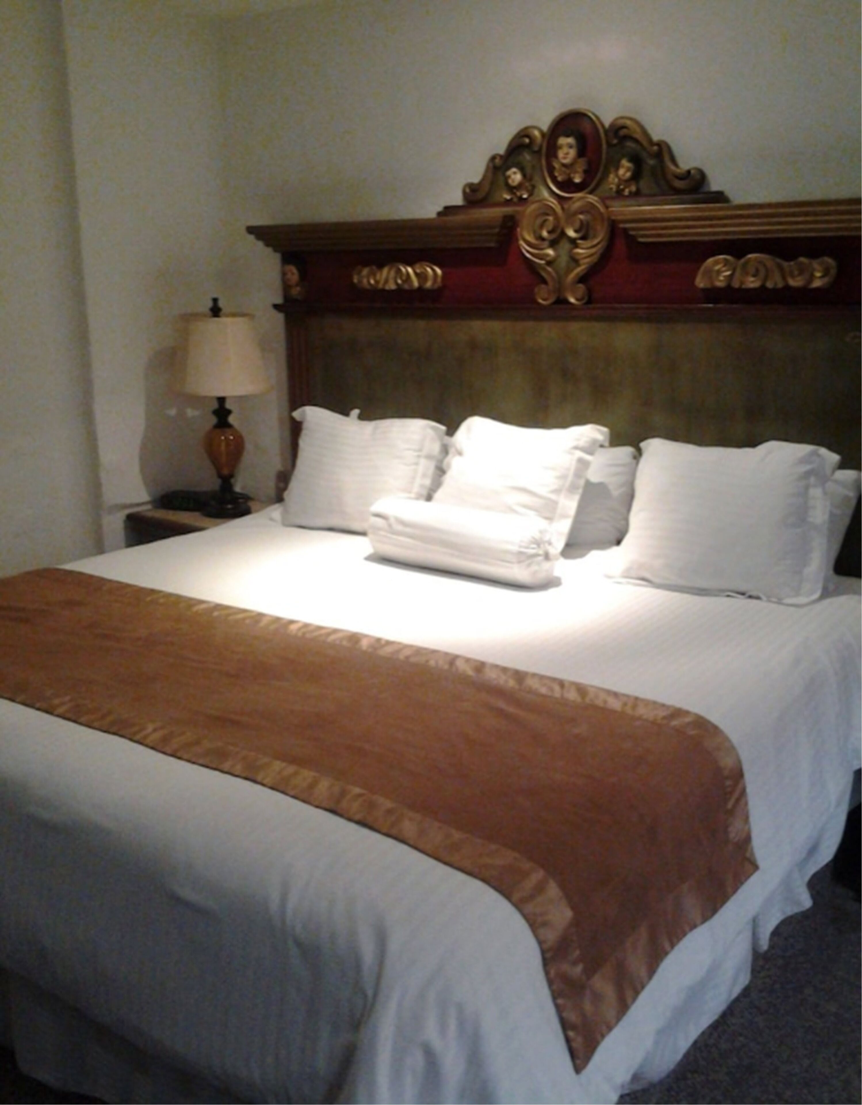 A neatly made bed with a brown bedspread and white pillows, a wooden headboard with decorative carvings, and a bedside table with a lamp.