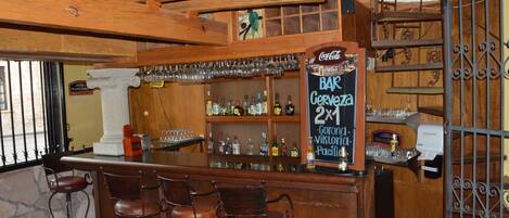 A bar area with a wooden counter, multiple stools, shelves with bottles, and a sign indicating a 2x1 beer offer.