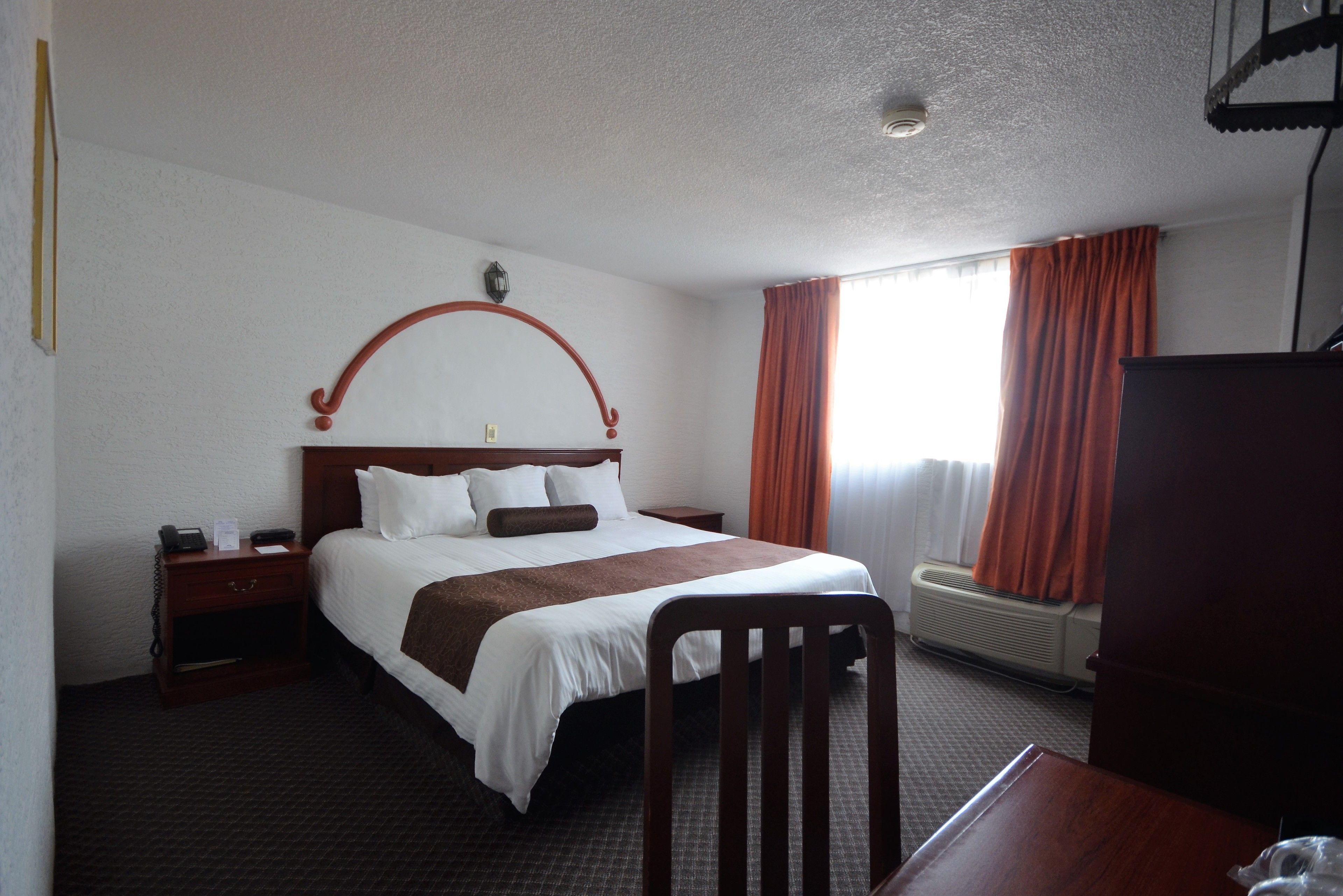 Habitación de hotel con una cama grande, cabecero de madera, mesita de noche con lámpara, escritorio con silla y ventana con cortinas.