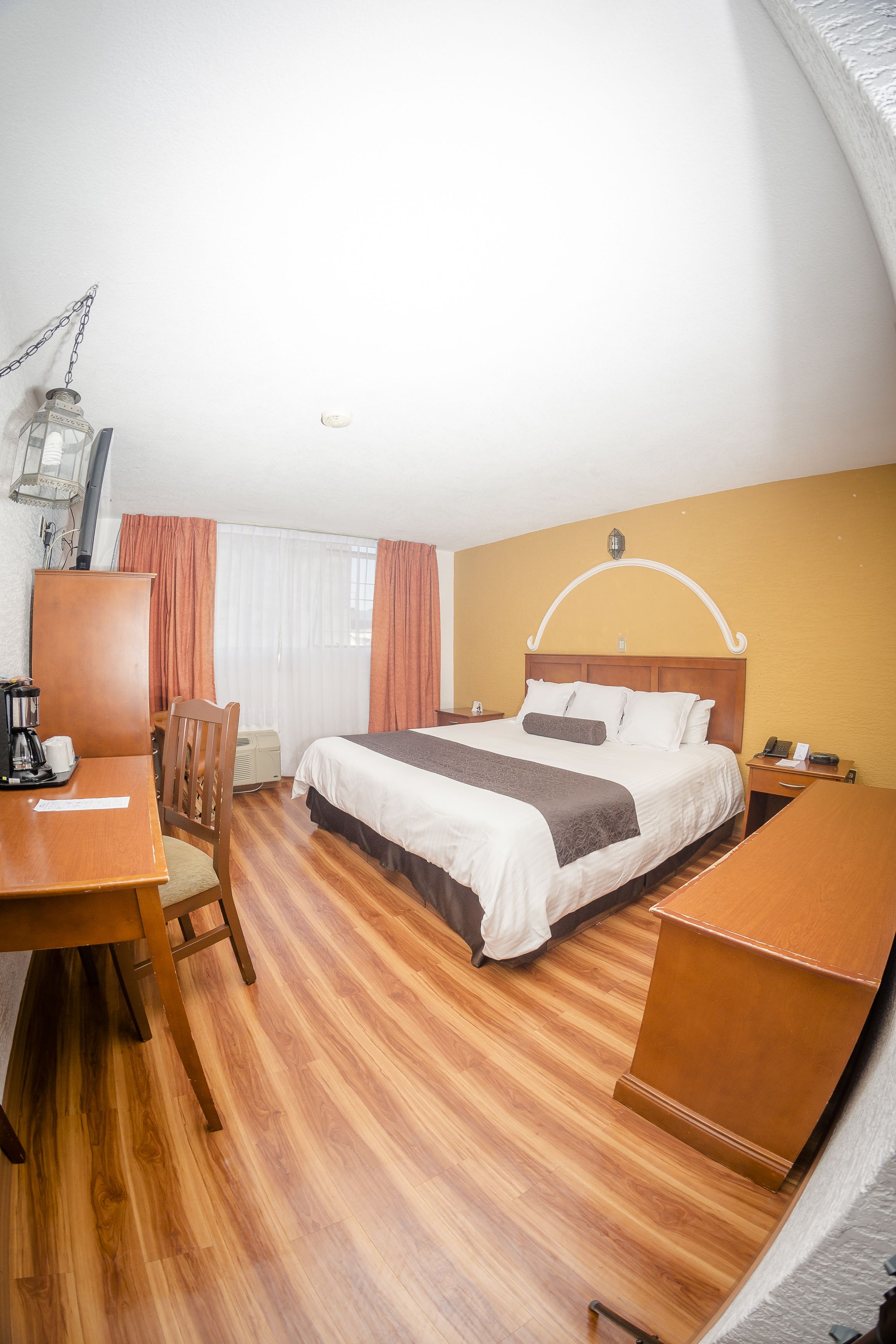Een hotelkamer met een houten vloer, een bed met wit en bruin beddengoed, een houten bureau met stoel en een nachtkastje met lamp.
