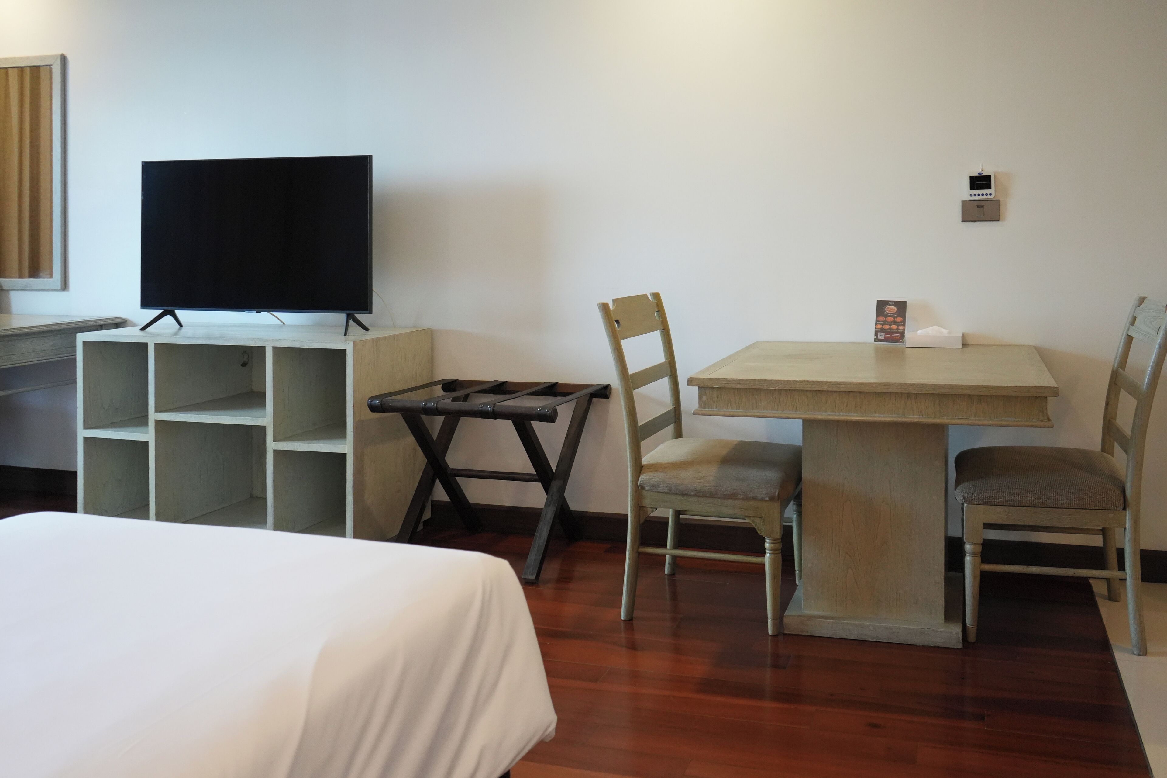 A hotel room with a bed, a desk with a television, a table with chairs, and a small shelf unit.