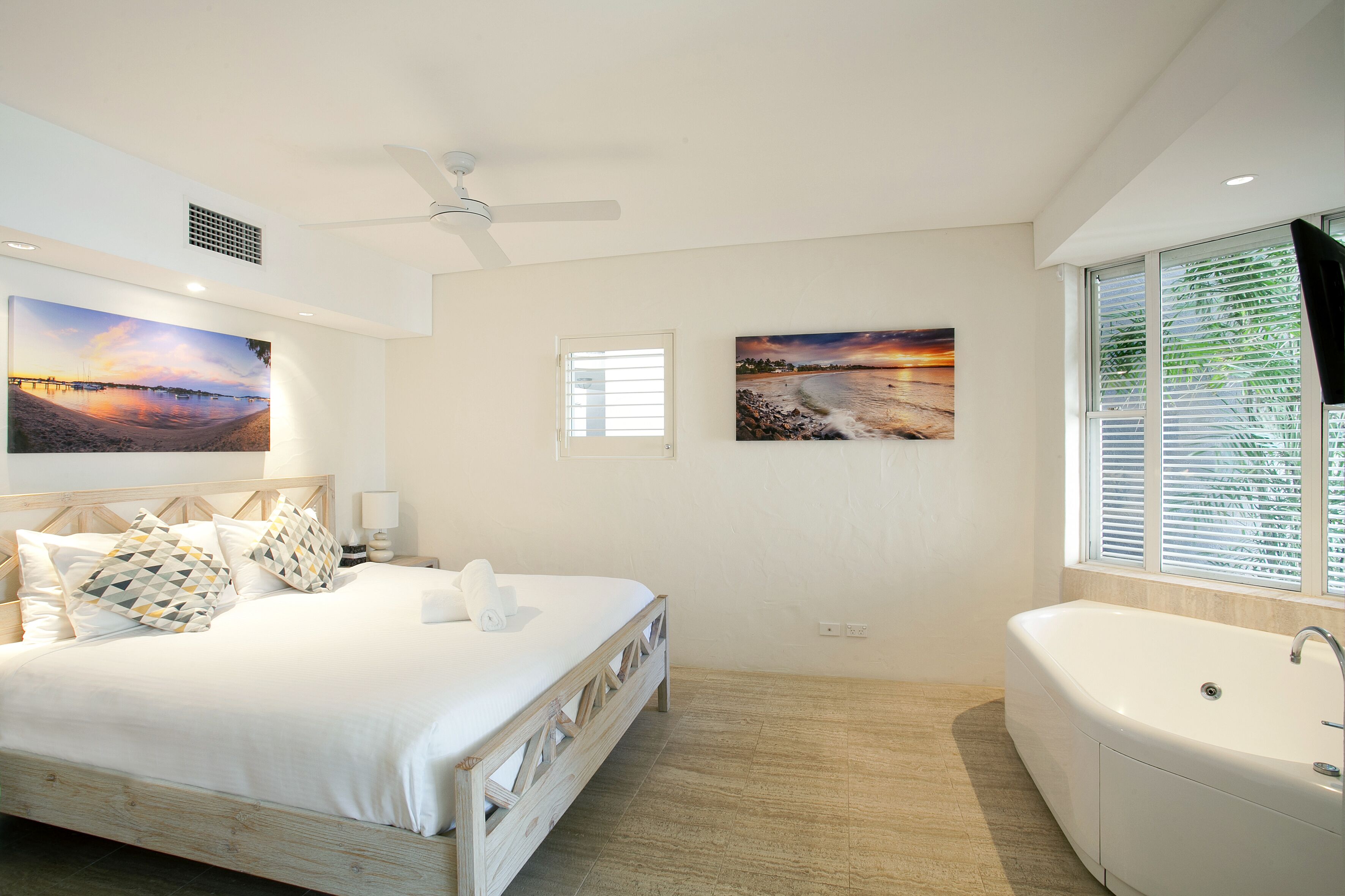 A bedroom with a bed, a large window with blinds, a ceiling fan, and two wall-mounted paintings.