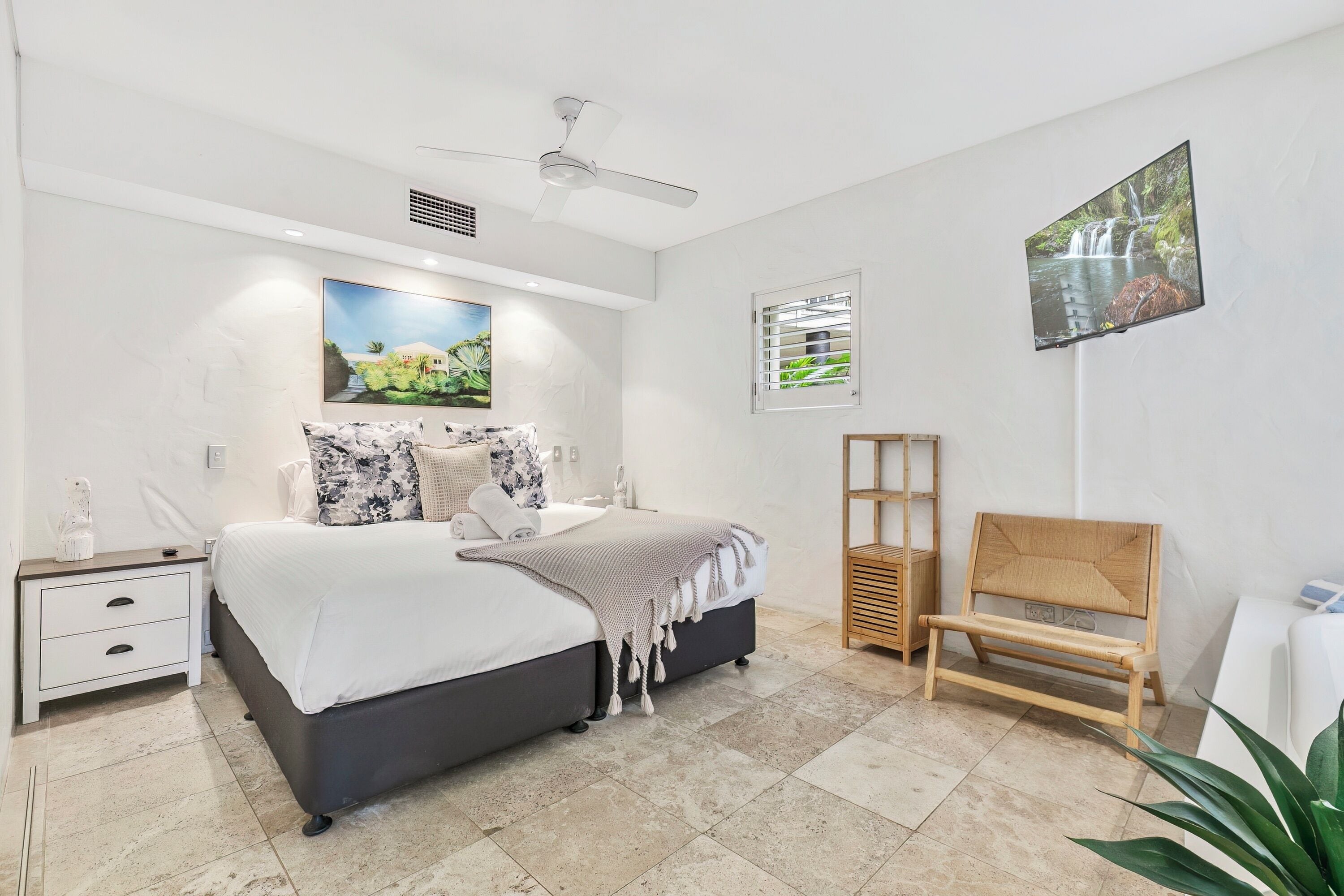 A modern bedroom with a bed, a nightstand, a bedside table, a ceiling fan, a TV mounted on the wall, and a window with blinds.