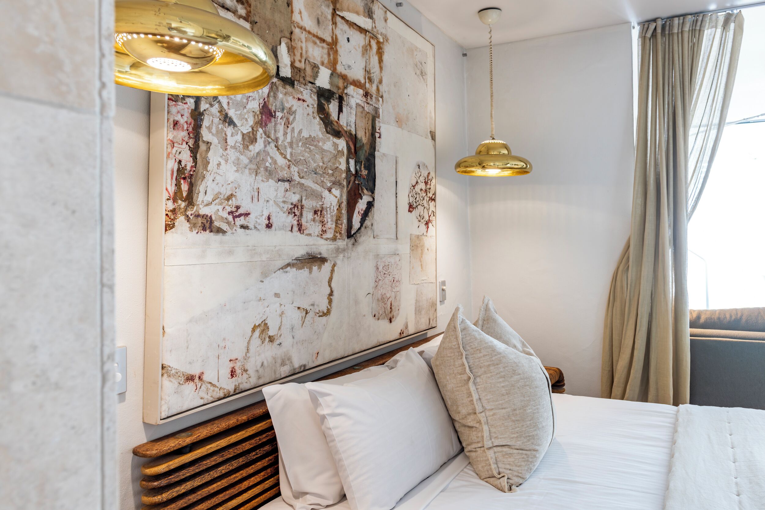 A hotel room with a large abstract painting on the wall, a bed with white linens, and a hanging gold pendant light.