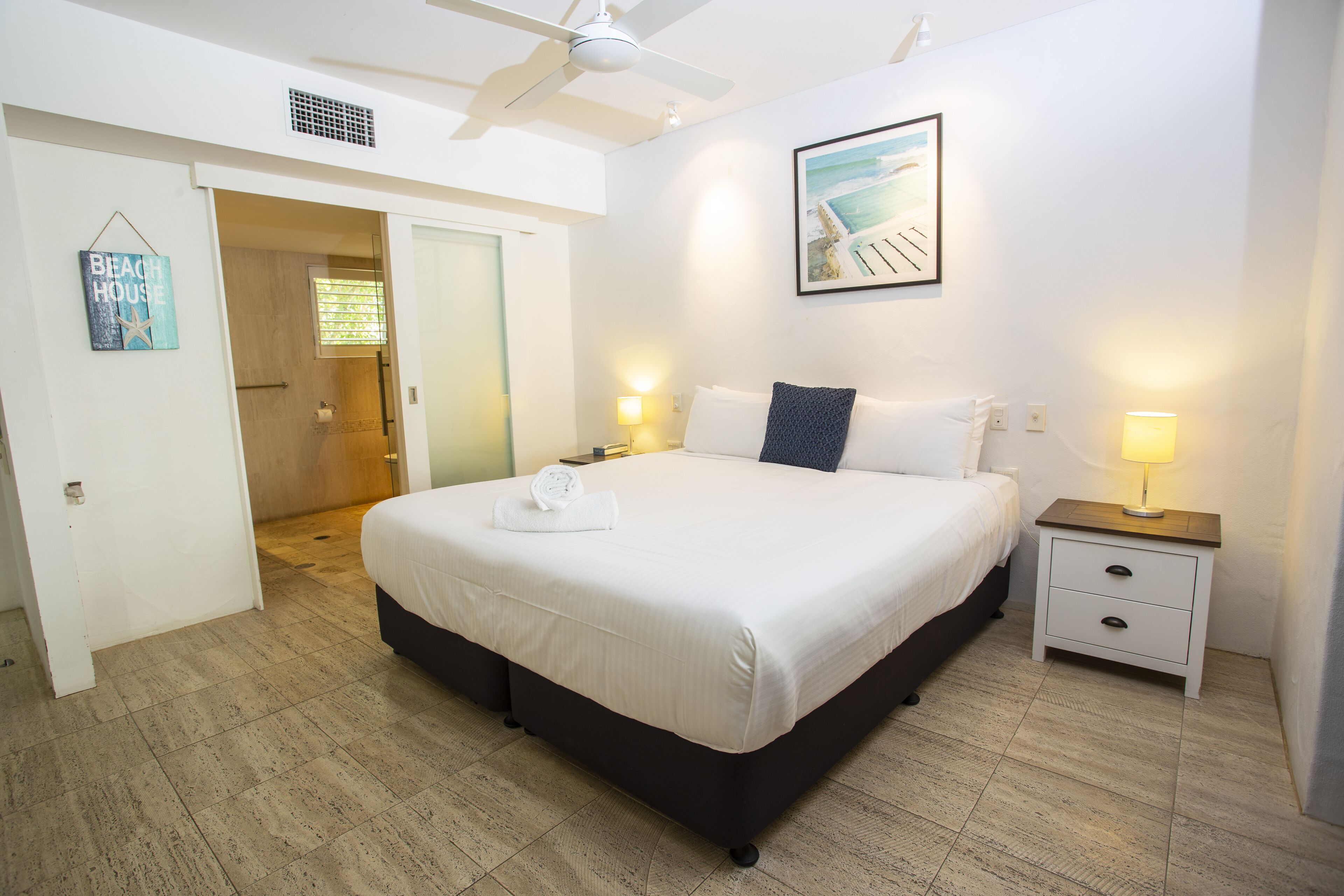 Quarto com cama, mesas de cabeceira, ventoinha no teto, porta e quadro fixado na parede.