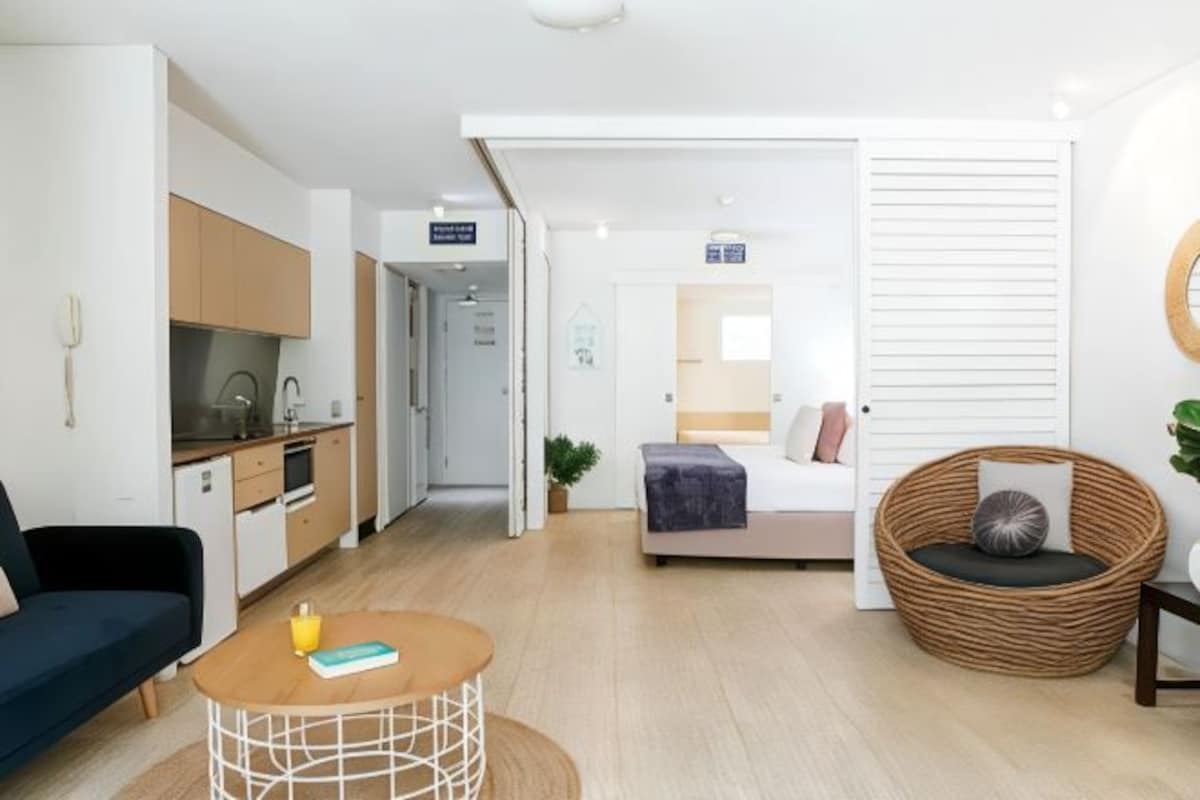 A modern living room with a sofa, a round coffee table, a wicker chair, and a kitchen area with cabinets and a sink.