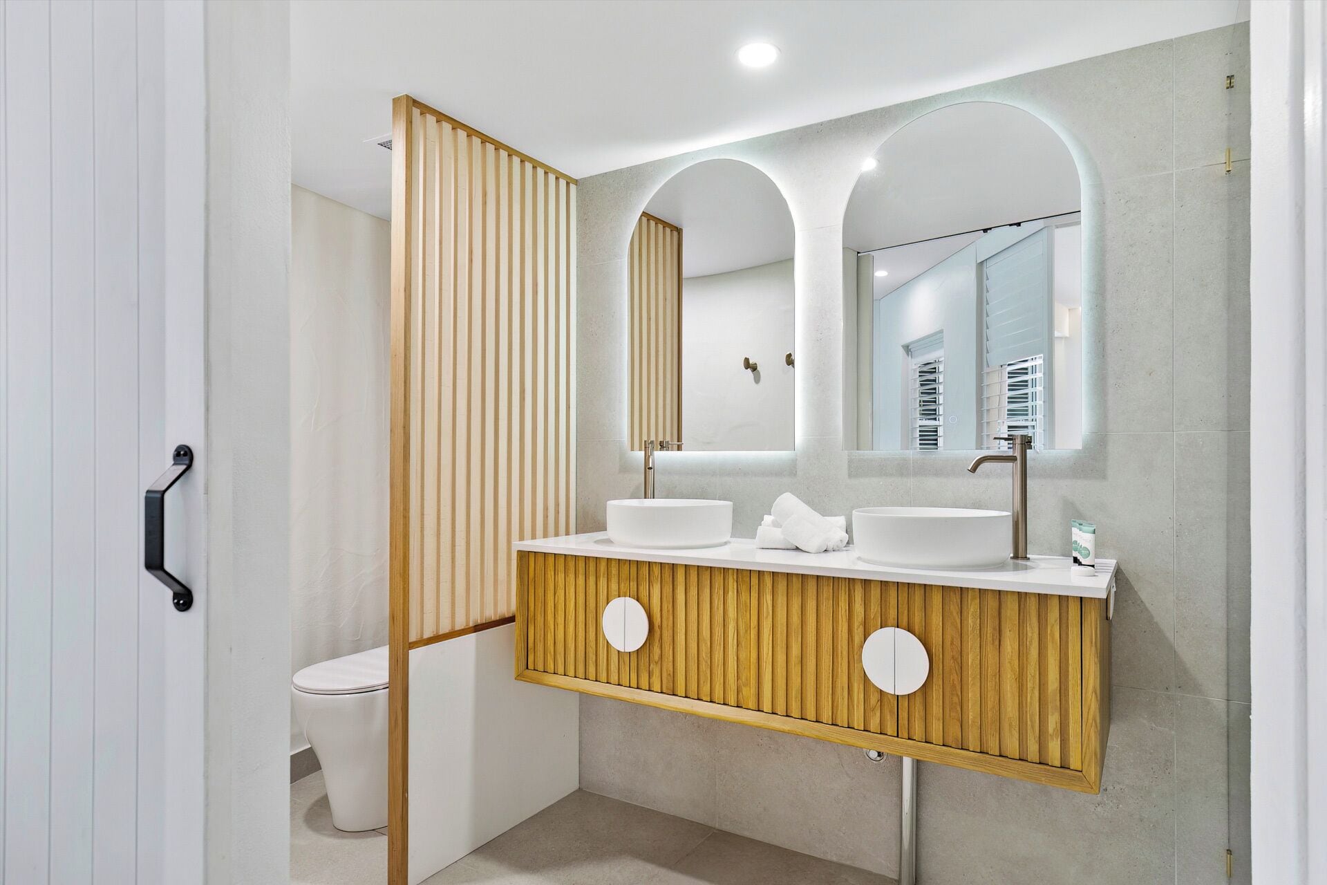 A modern bathroom with a wooden vanity, two sinks, and a large mirror with an arched top.