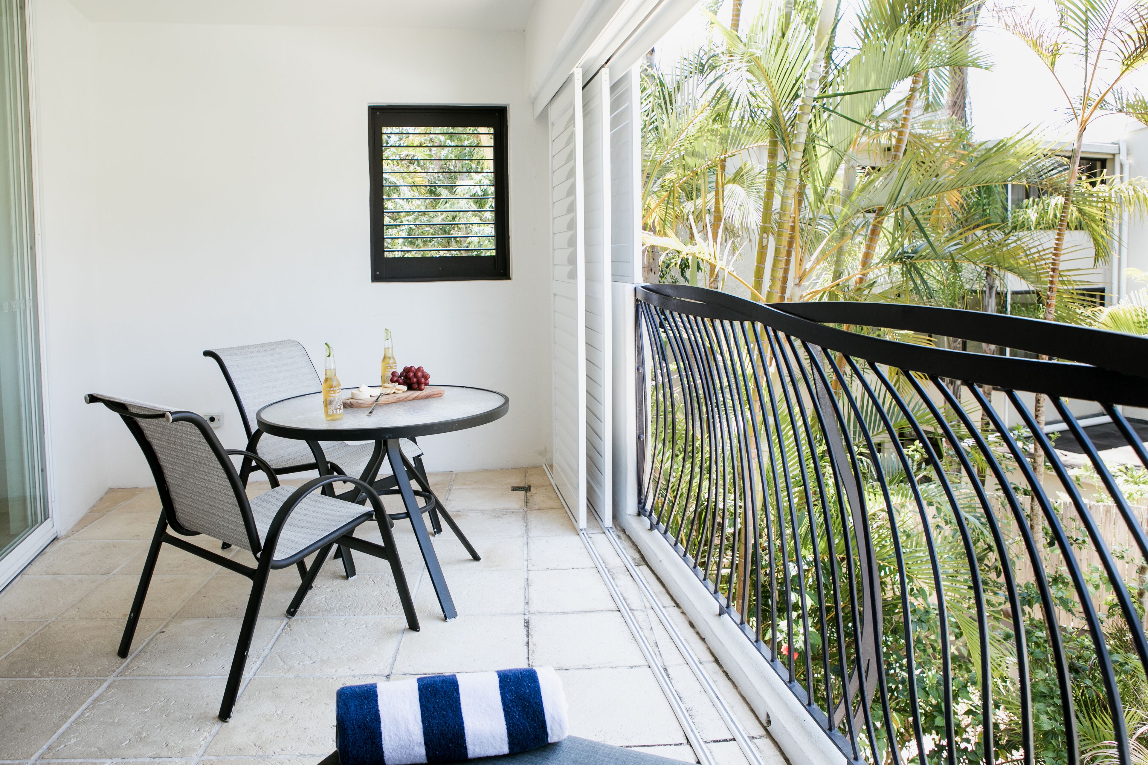 A balcony with a table and chairs, a window with blinds, and a view of a garden with palm trees.