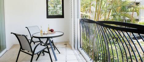 A balcony with a table and chairs, a window with blinds, and a view of a garden with palm trees.