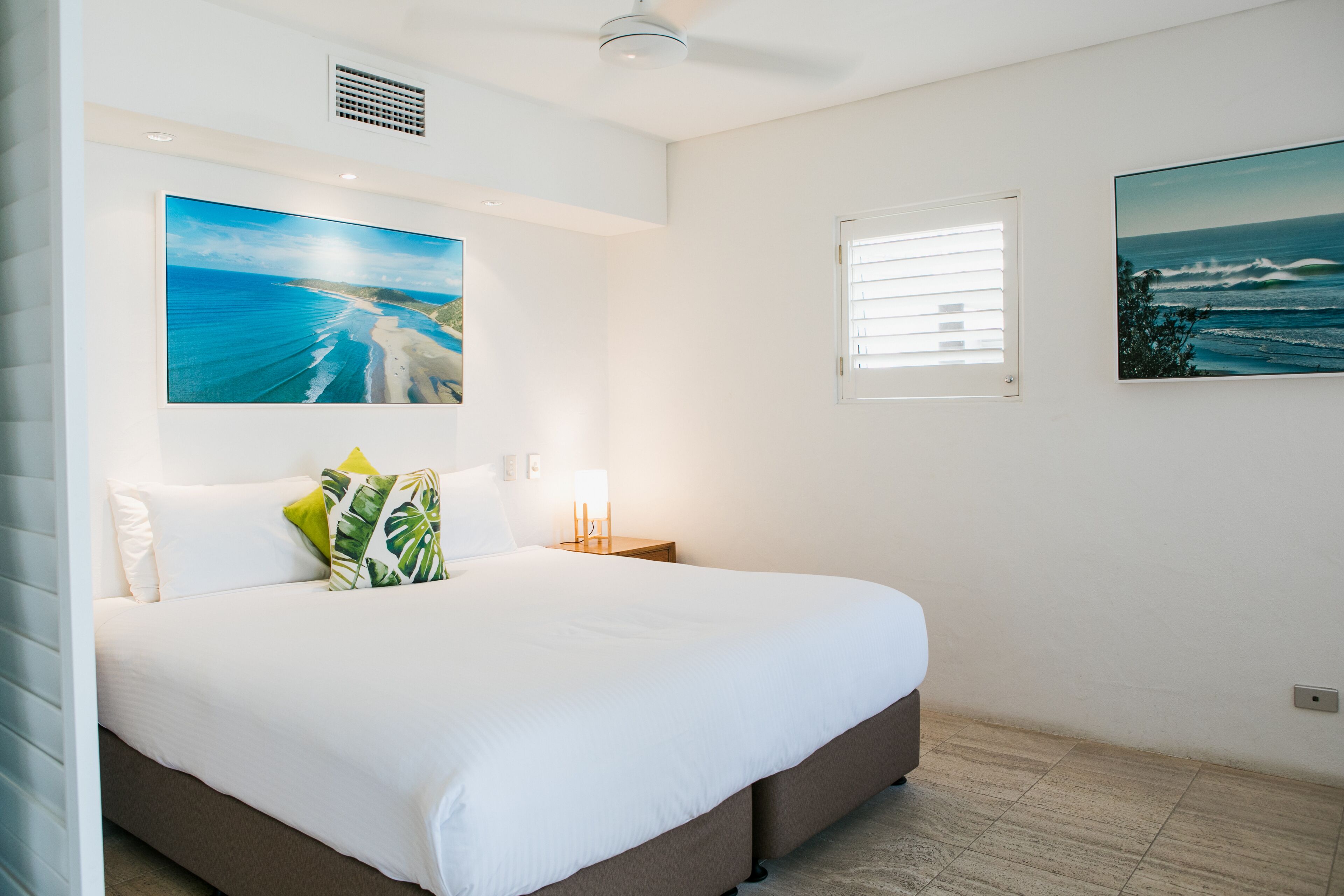 A bedroom with a large bed, a nightstand with a lamp, and two wall-mounted paintings of the ocean.