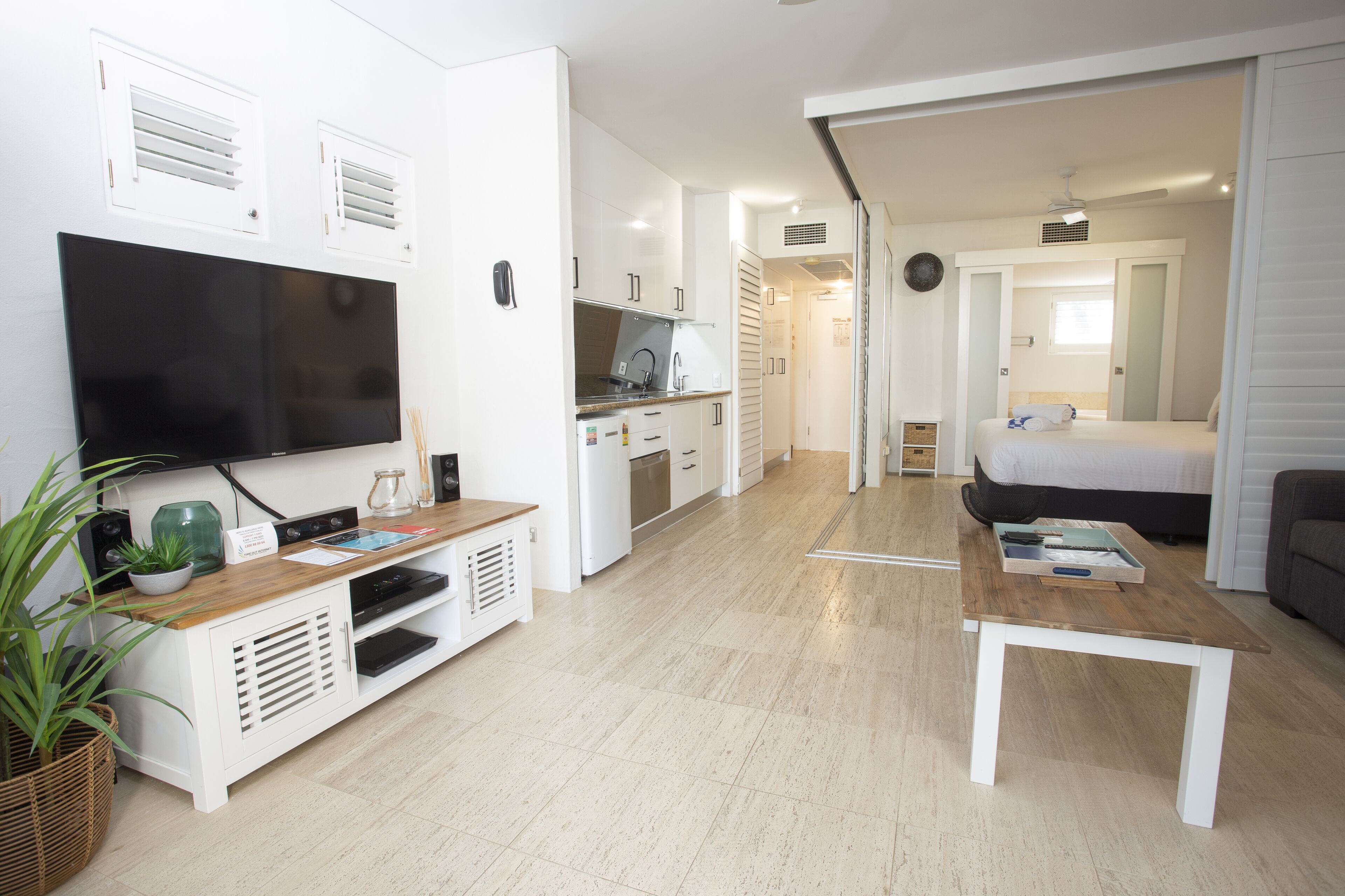A modern living room with a flat-screen TV, a coffee table, and a sofa. There is a kitchen area with cabinets and a microwave. The room has a wooden floor and white walls.