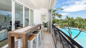 A balcony with a wooden table and bar stools, overlooking a swimming pool and palm trees.