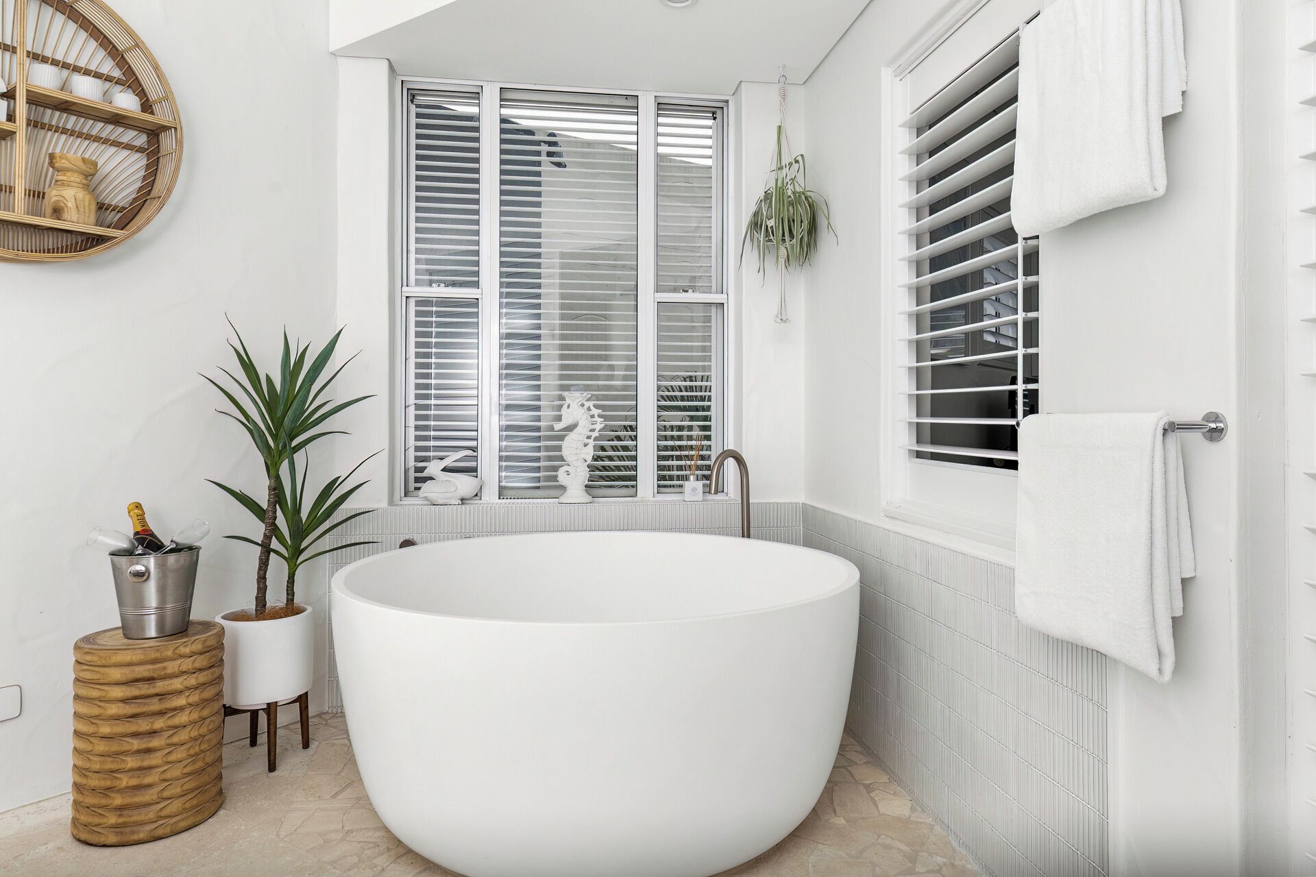 A modern bathroom with a freestanding bathtub, a window with blinds, a potted plant, and a towel rack.