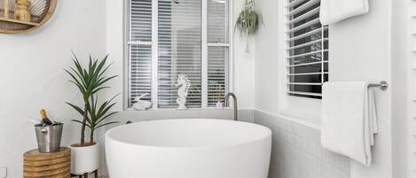 A modern bathroom with a freestanding bathtub, a window with blinds, a potted plant, and a towel rack.