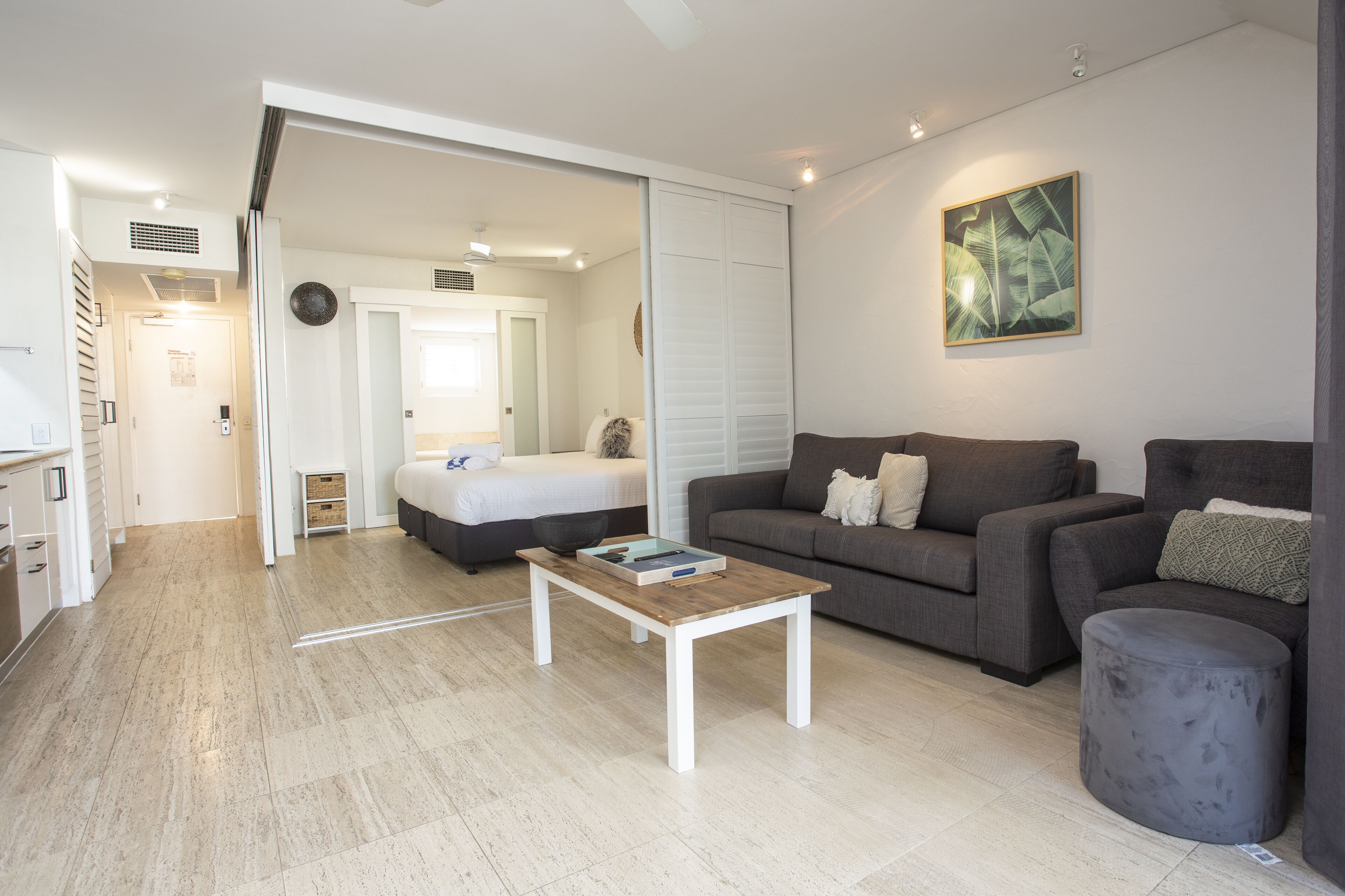 A modern living room with a sofa, a coffee table, and a grey ottoman. There is a bed in the background and a door leading to another room.