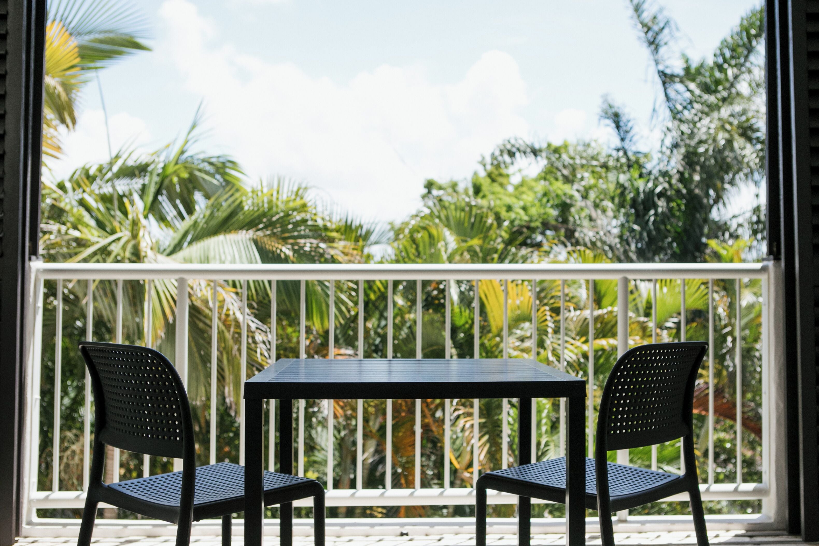 A balcony with two black chairs and a table, overlooking a tropical garden with various plants and trees.
