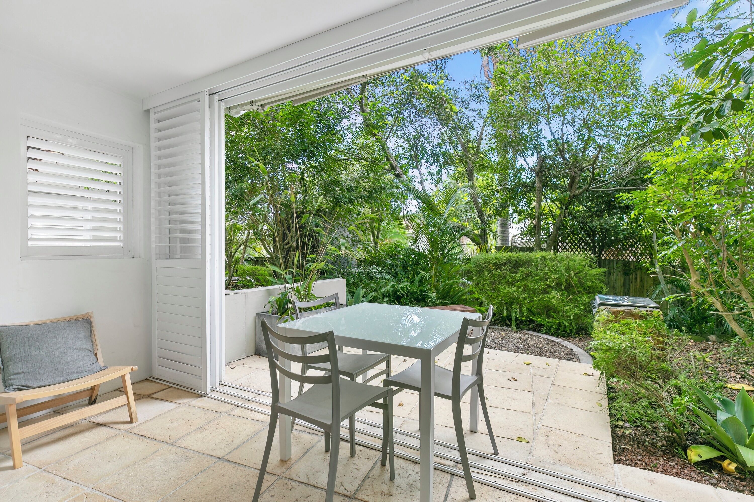 A patio with a glass table and chairs, a bench, and a view of a lush garden.